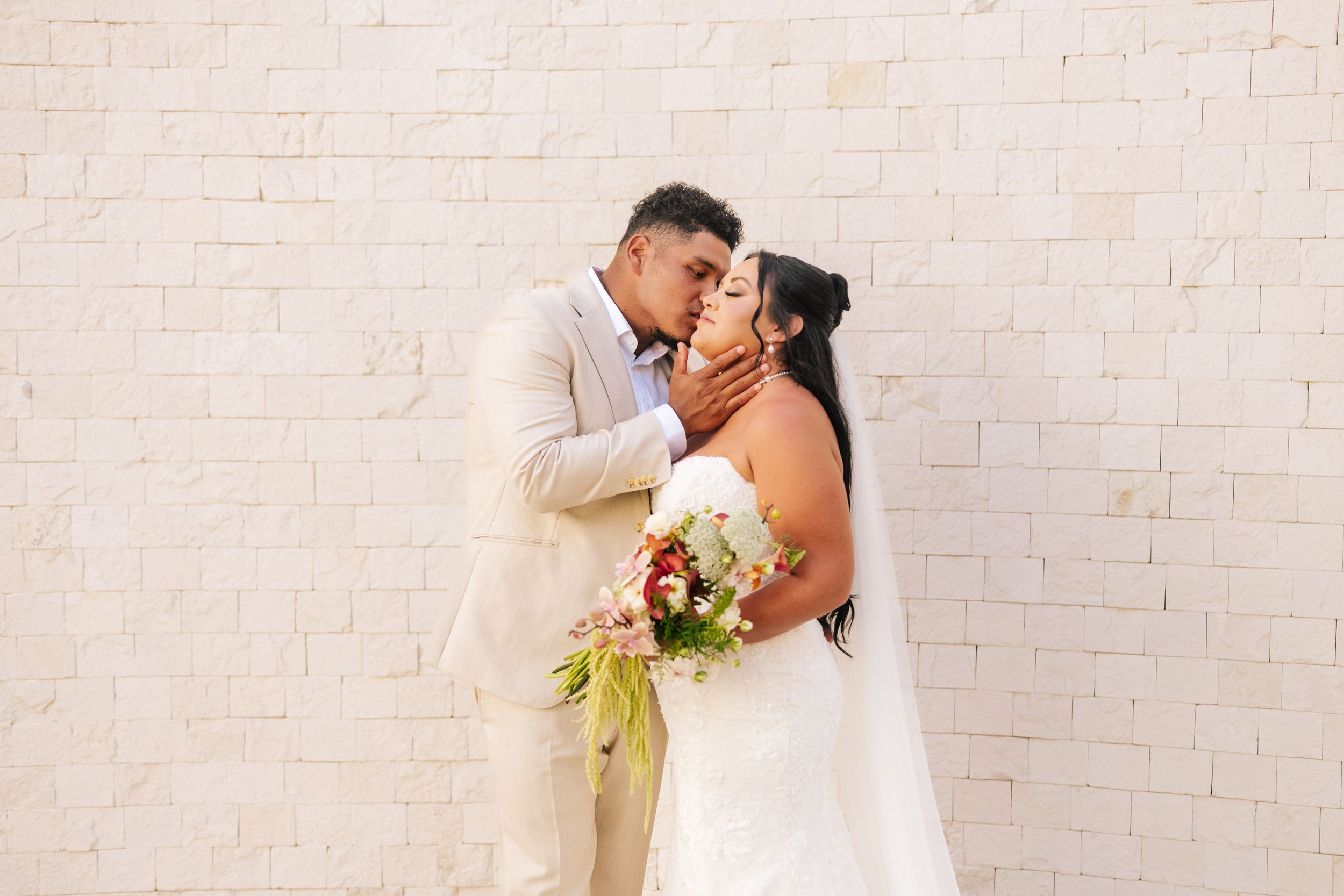 Bride in a white gown holding a colorful bouquet embraces her groom in a tan suit for a romantic kiss against a white brick wall.