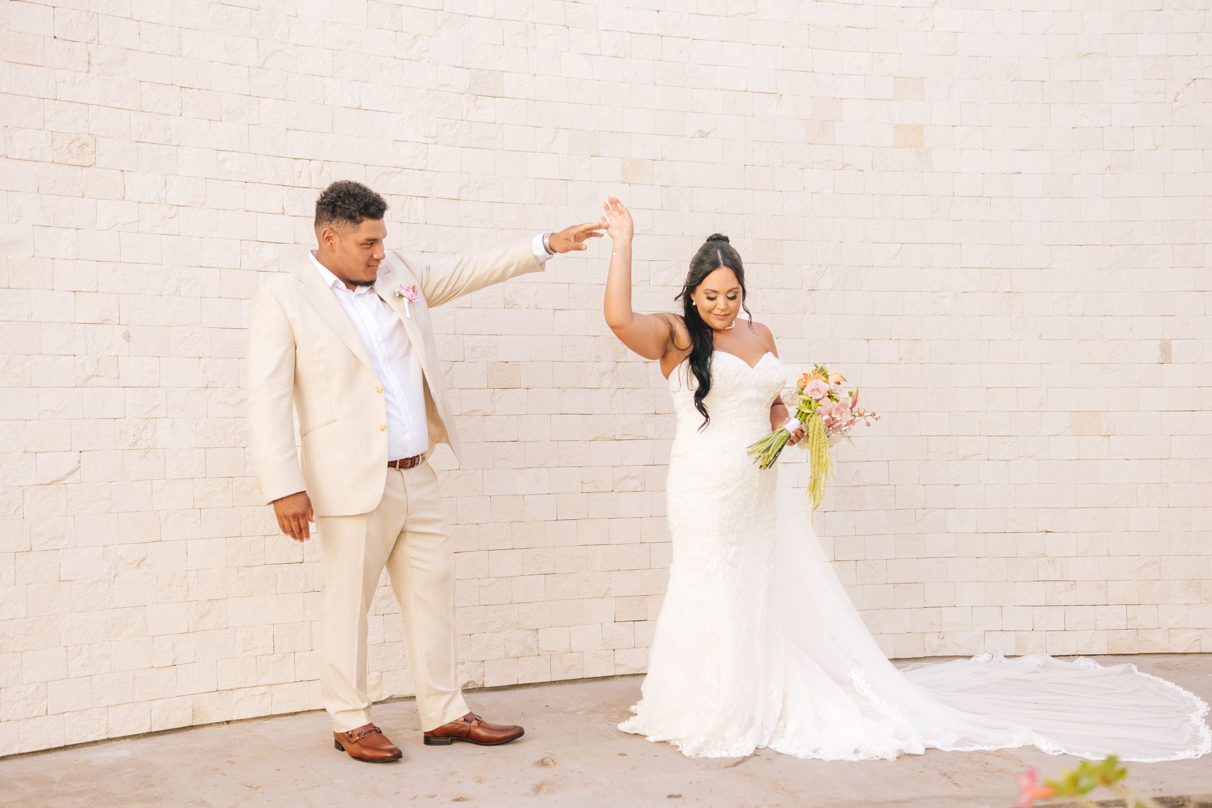Bride in a white gown with a long train twirls with a bouquet as the groom in a cream suit reaches toward her against a white brick wall.