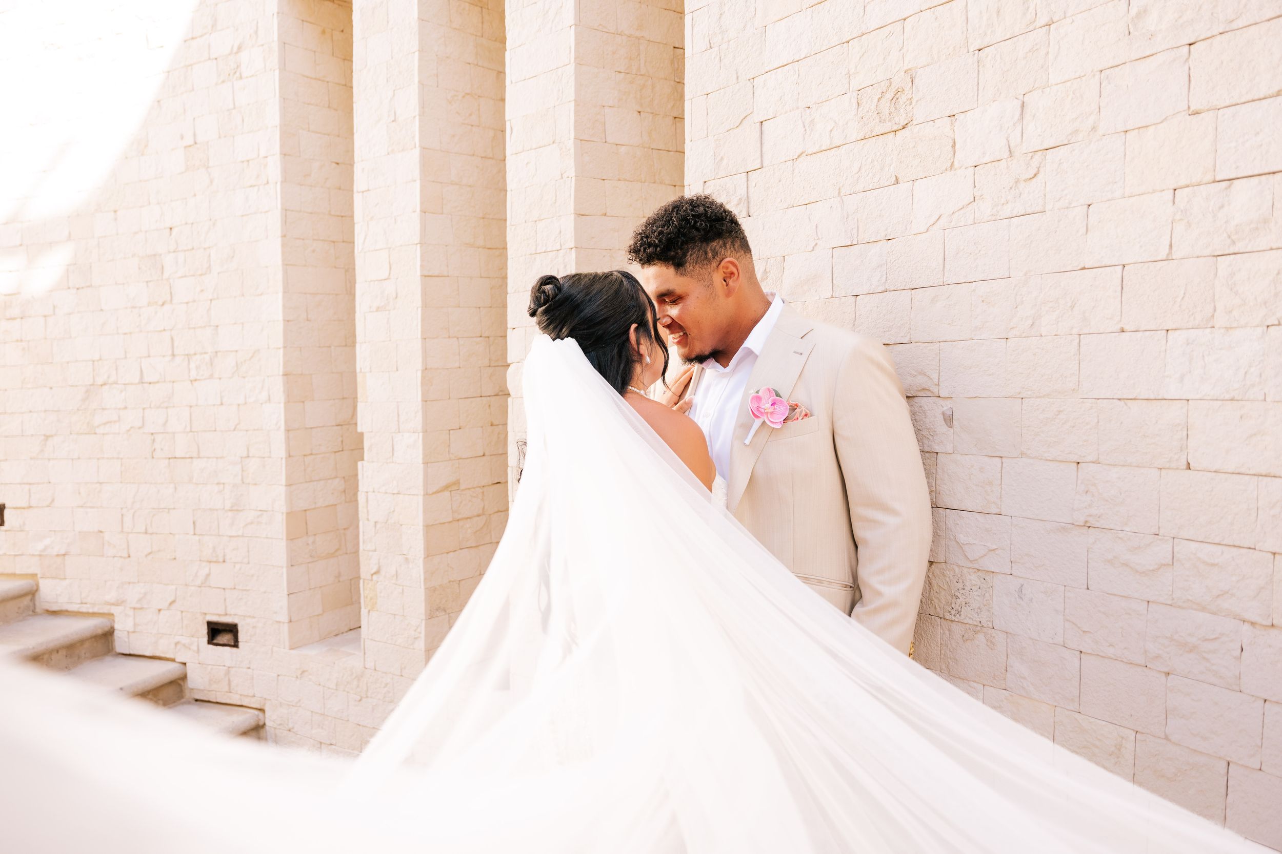 A bride and groom share an intimate moment against a sunlit stone wall, her long wedding veil flowing dramatically around them.