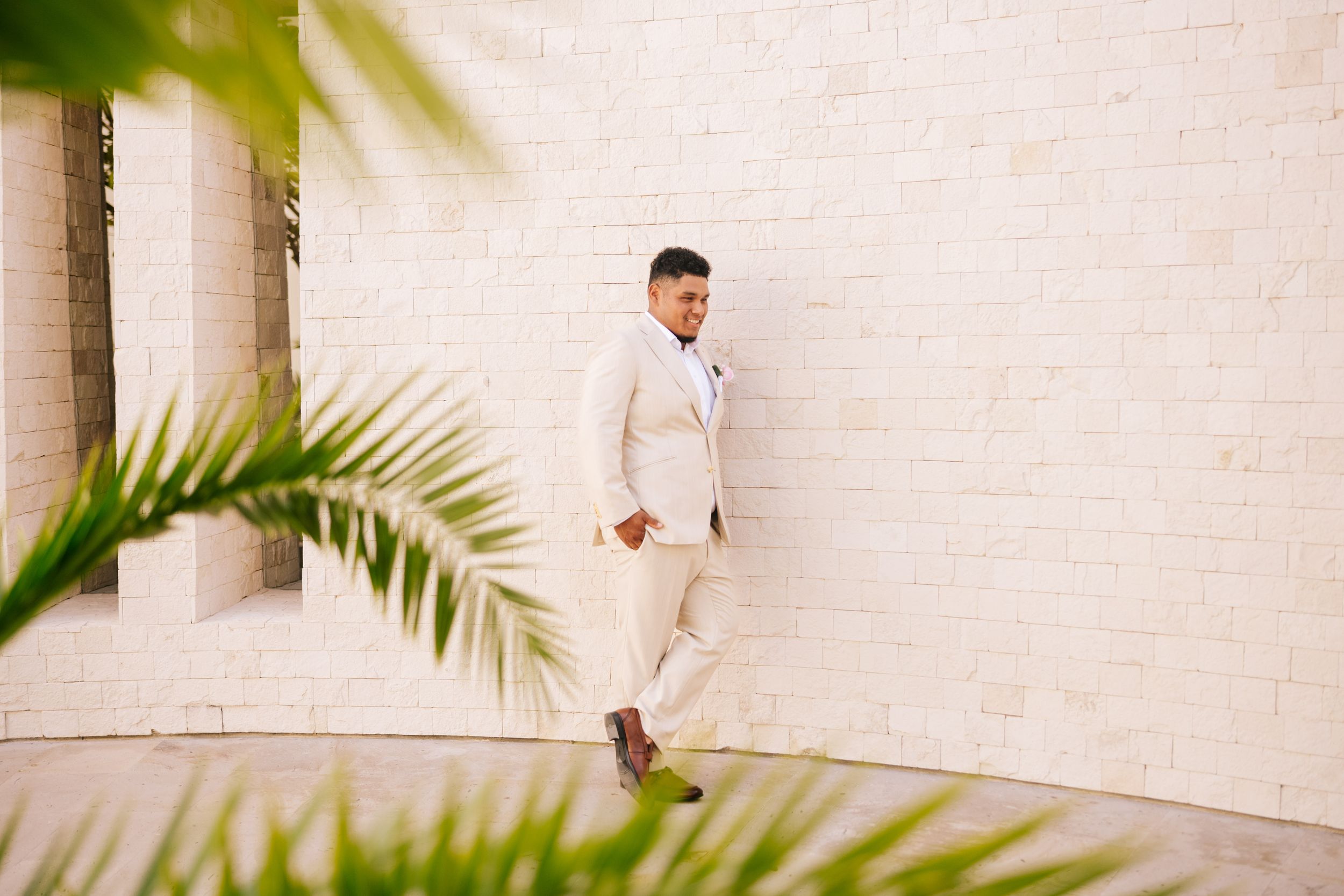 Man in a cream linen suit and brown shoes walks along a white brick wall, framed by green palm fronds in the foreground.