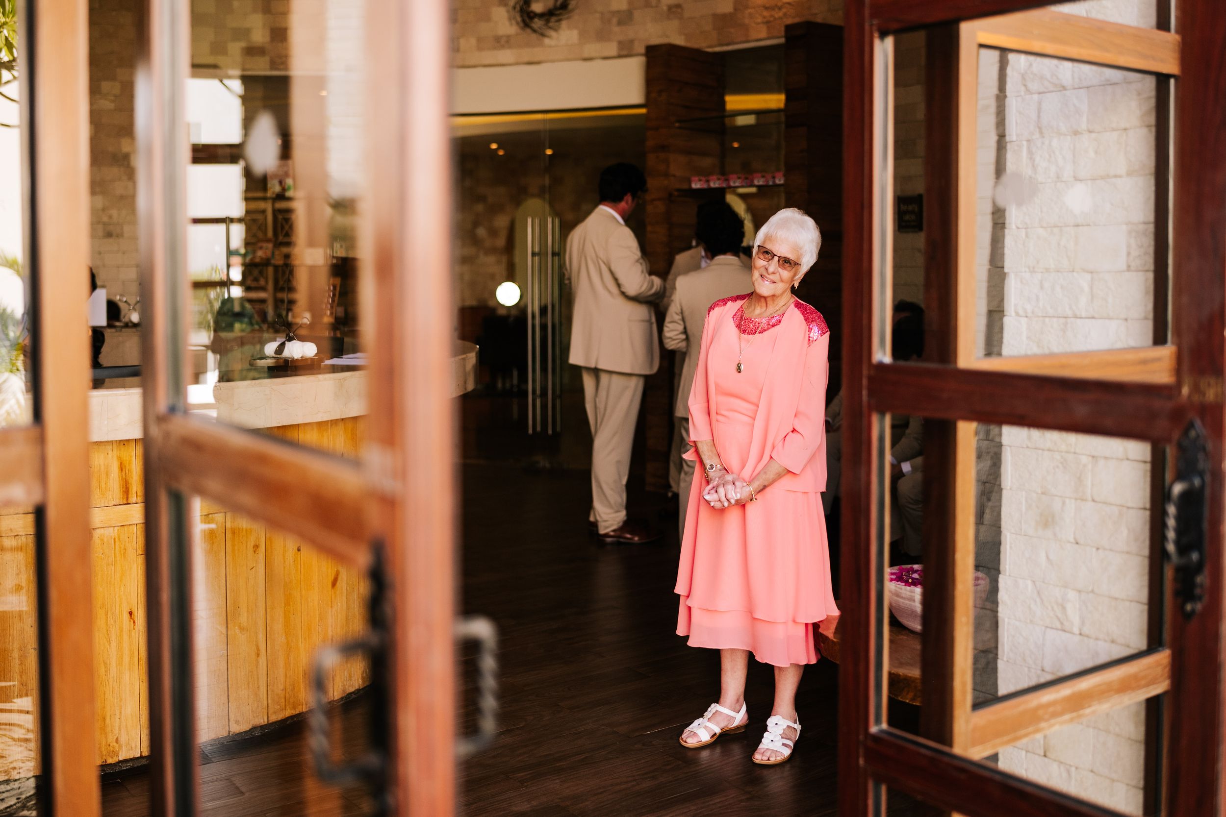 Elderly woman in a coral pink dress and sandals smiles in a wooden doorway while two people stand in a warm, modern interior behind her.