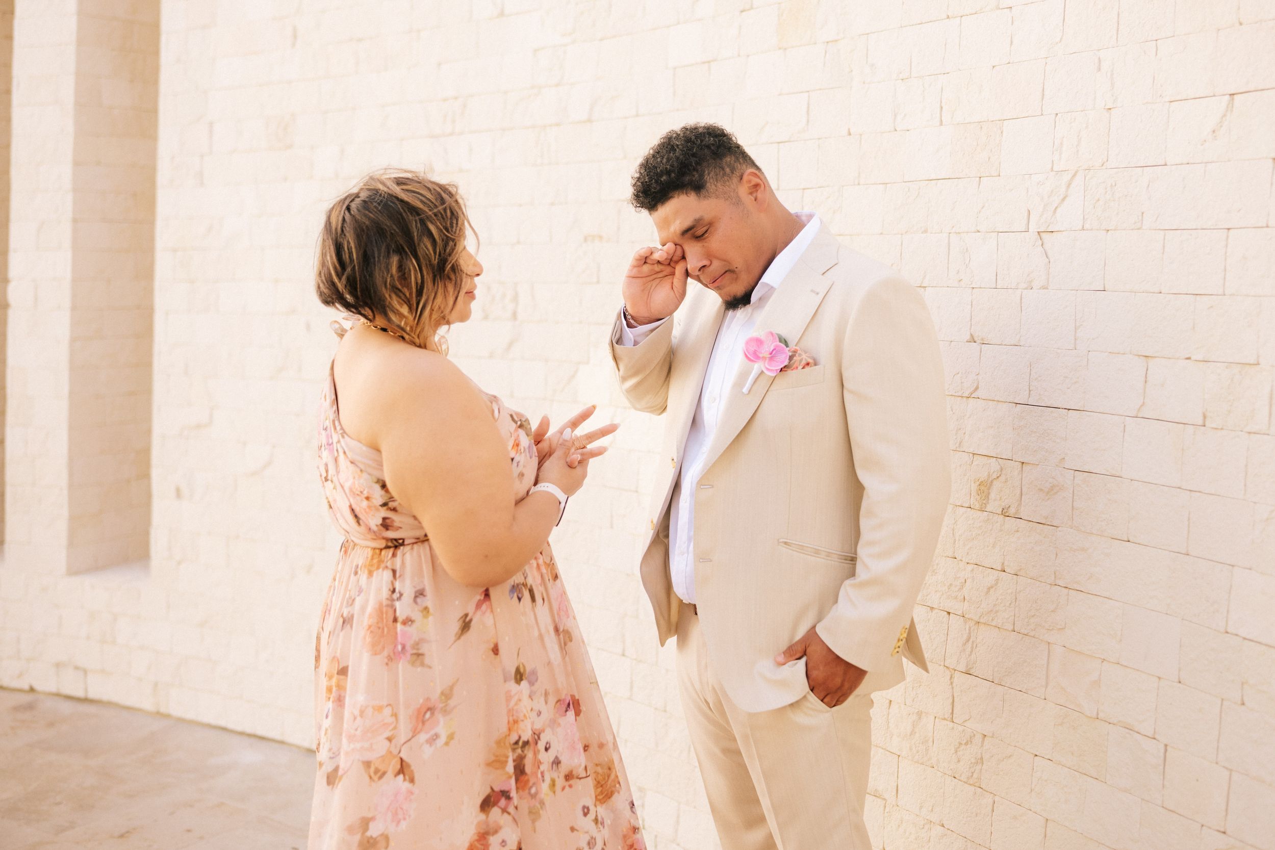 Emotional bride in a floral blush dress and groom in a cream suit with pink boutonniere wiping tears before a white brick wall.