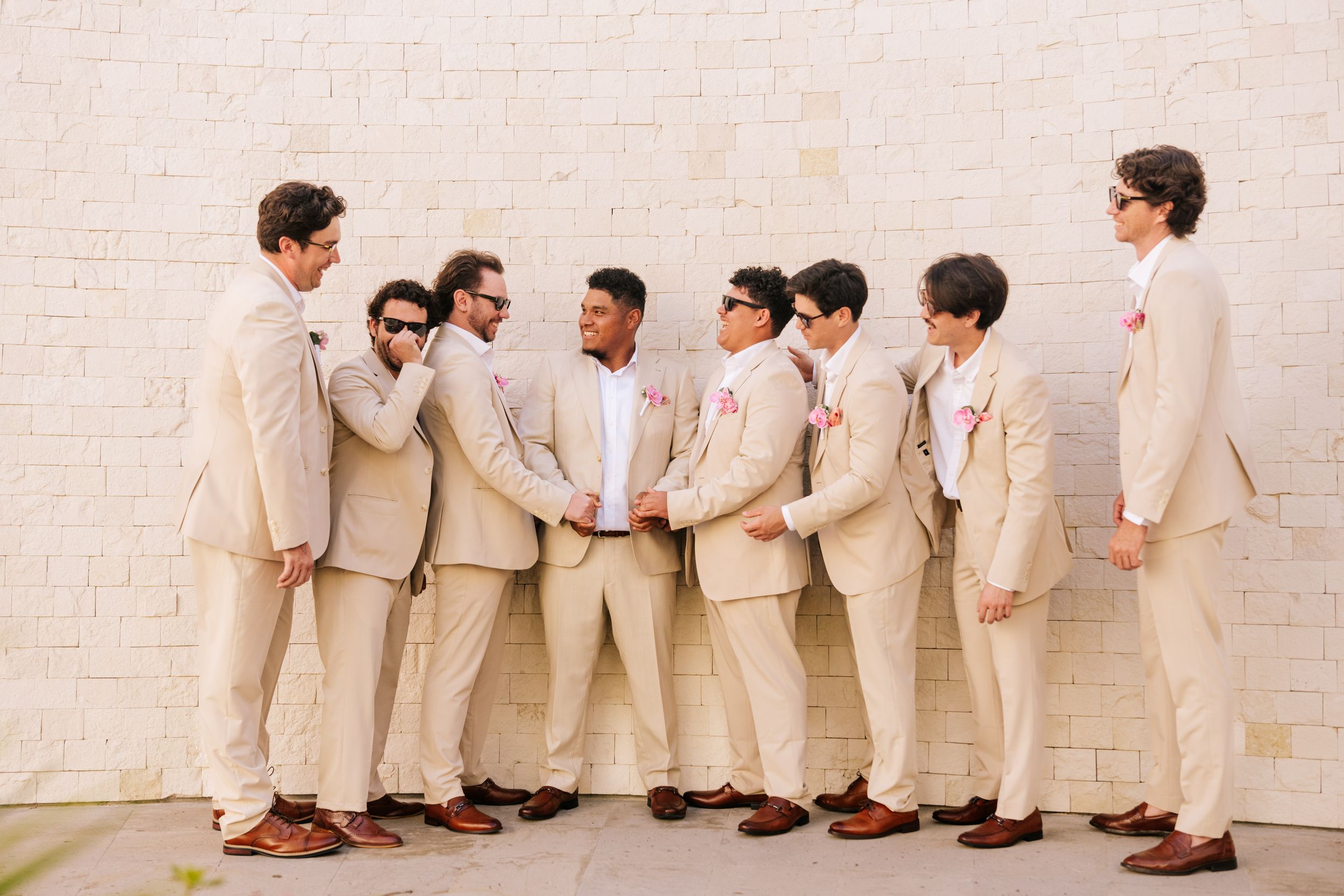 Groomsmen in matching tan suits and brown leather shoes laugh and pose together in a casual group against a white brick wall.