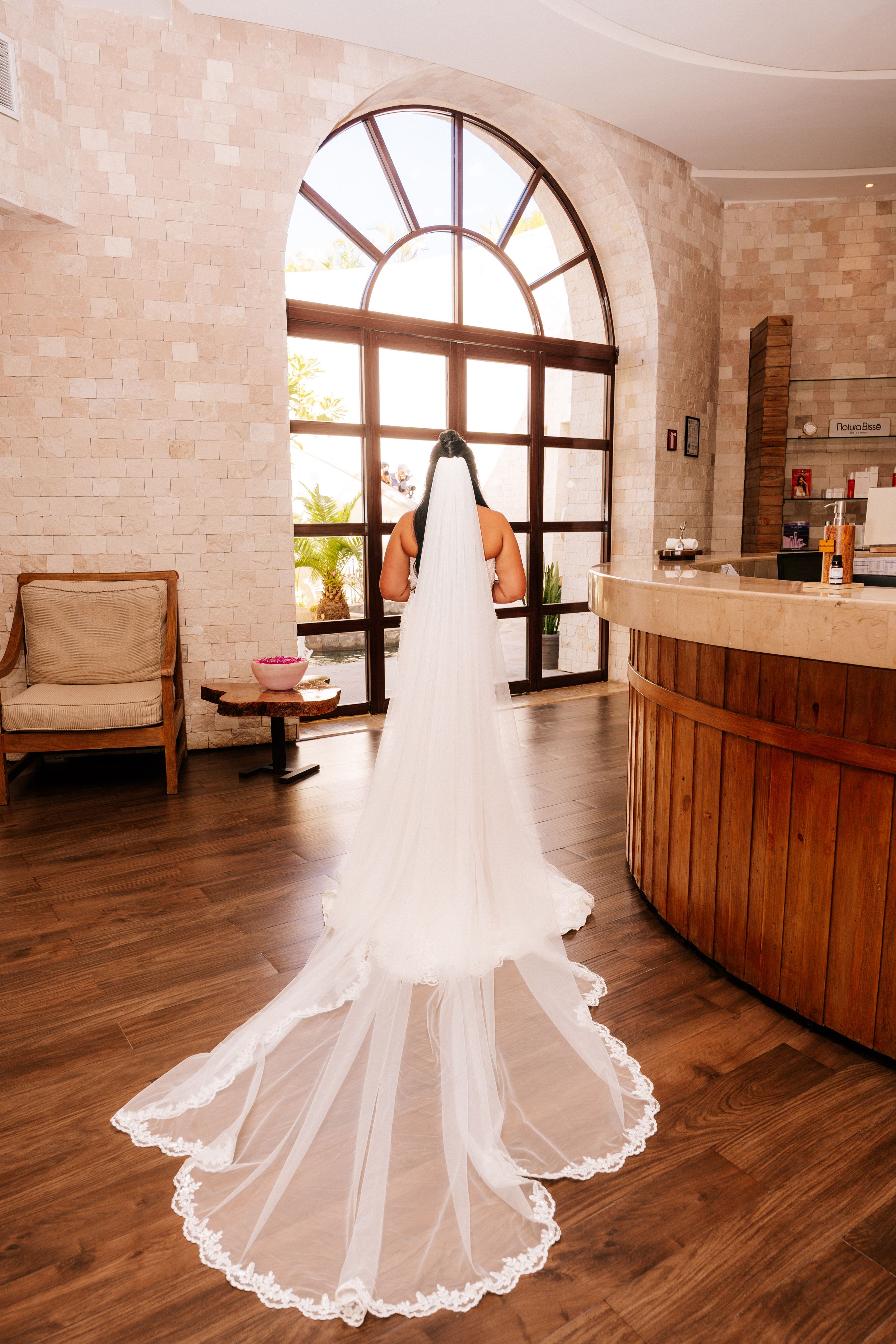 Bride seen from behind in a long lace-trimmed cathedral veil standing on a wood floor beside a curved counter and arched window.