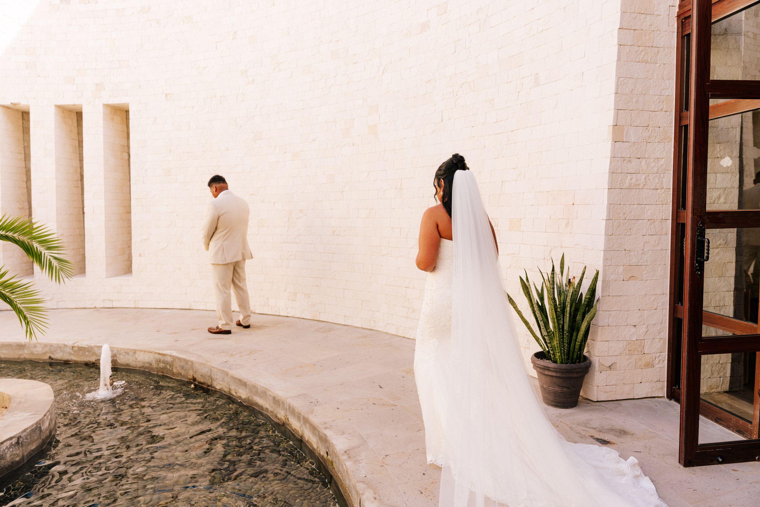 Bride in a long white veil and gown stands near a courtyard fountain as the groom in a tan suit waits with his back turned for a first look.