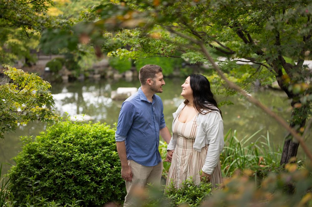 Smiling couple in casual clothes stand close together in a lush garden beside a calm pond, framed by green foliage and trees.