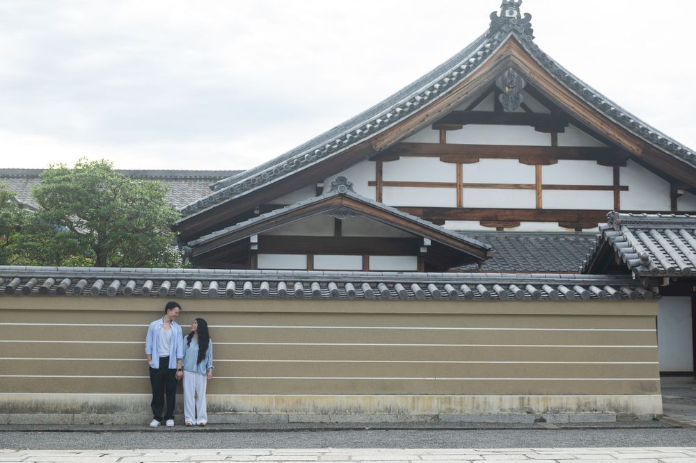 Couple standing side by side in front of a traditional Japanese temple with a curved tiled roof and a long striped earthen wall.