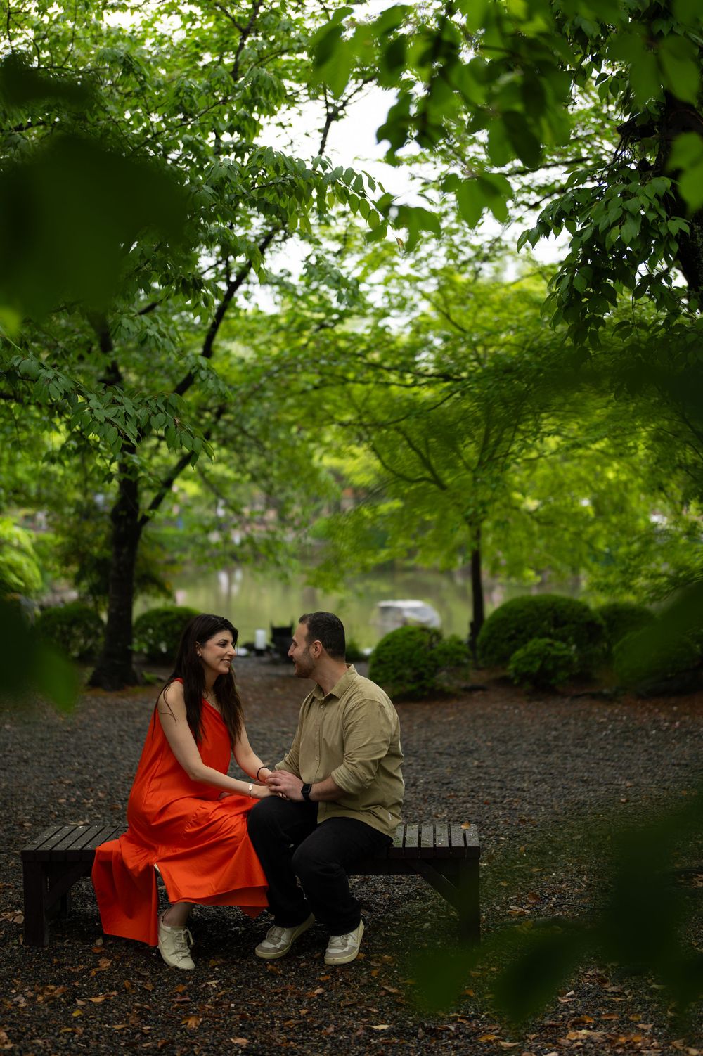 Couple in an orange dress and olive shirt sit holding hands on a wooden park bench, surrounded by lush green trees near a calm pond.