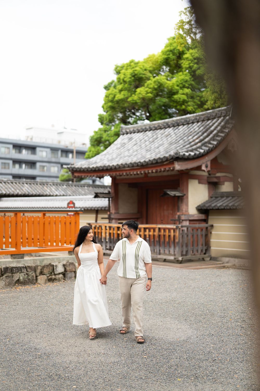 Couple holding hands and walking past a traditional Japanese temple building with a tiled roof and orange fence on a gravel path.