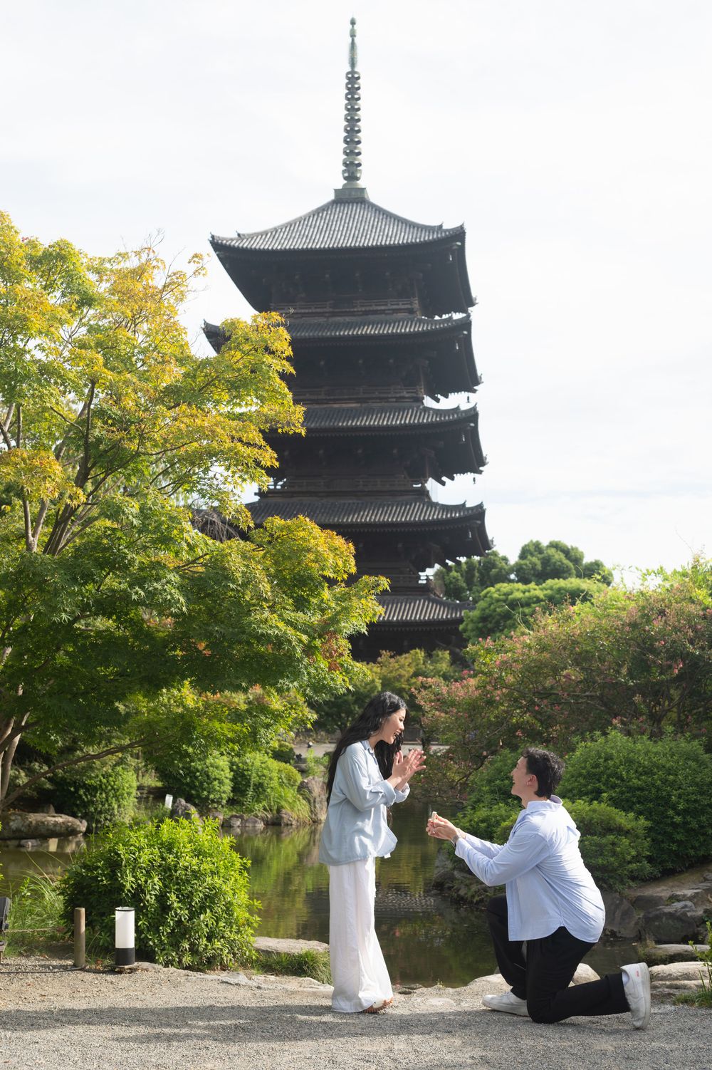 Man kneeling on one knee proposing to a woman in front of a tall Japanese pagoda surrounded by green trees and a calm pond.