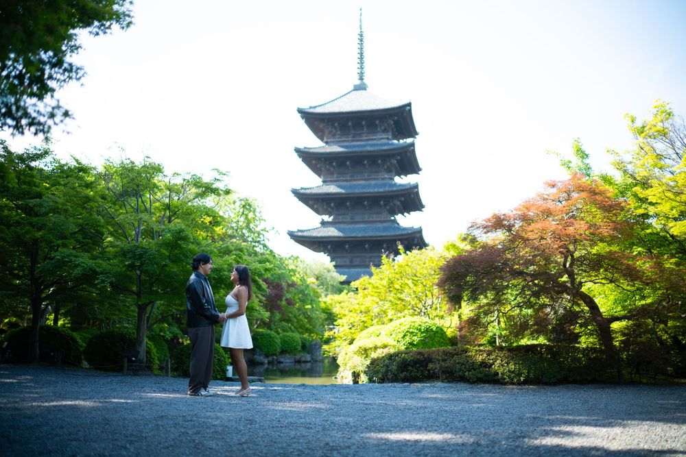 Couple in formal attire stands hand in hand on a gravel path before a tall five-story pagoda surrounded by lush green and autumn trees.