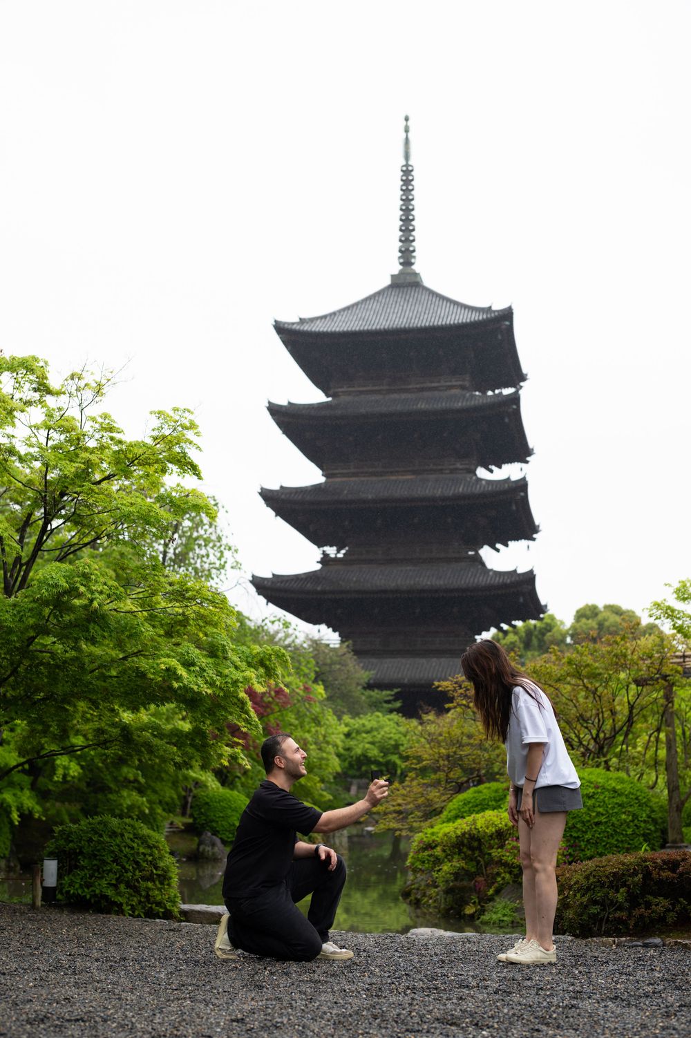 Man kneeling on one knee in front of a woman in a garden, with a tall five-story Japanese pagoda rising behind lush green trees.