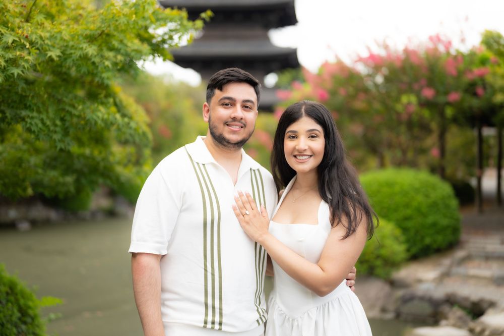 Smiling couple embracing outdoors in a lush garden with a pagoda, pink flowering trees, and a calm stream in the background.
