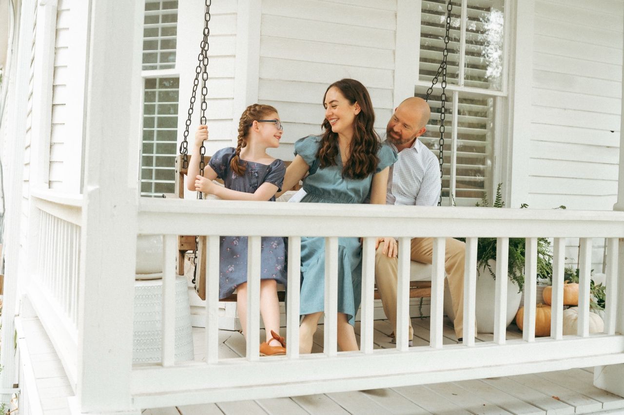 Family of three smiling together on a white porch swing, enjoying a relaxed moment outdoors in McKinney, TX