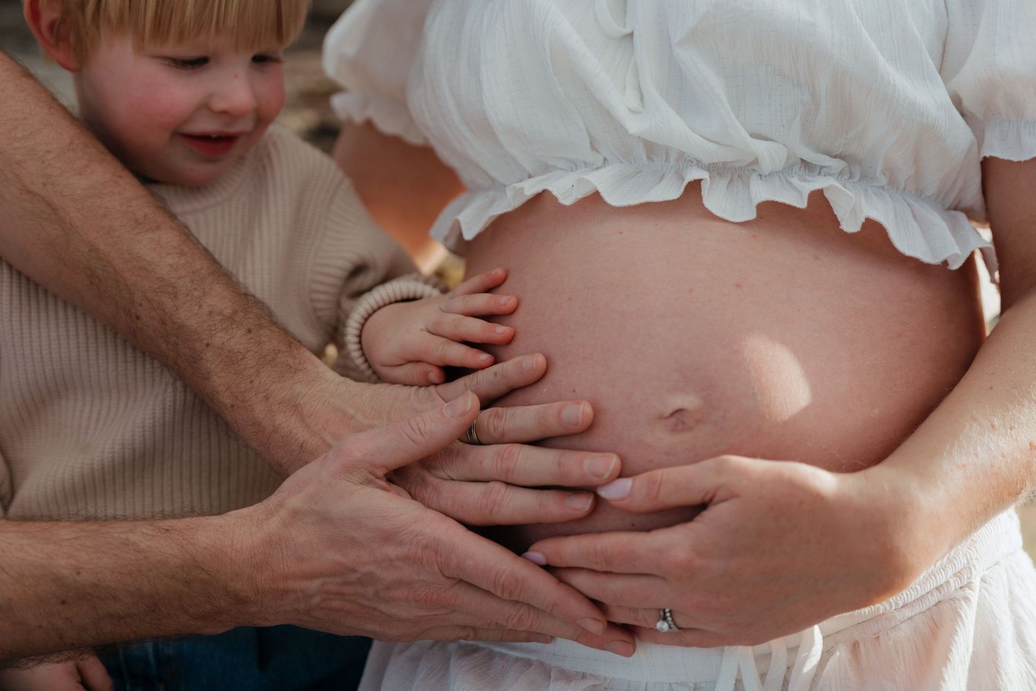 A pregnant woman's bare belly being gently touched by a man and young toddler child during a maternity photo session.