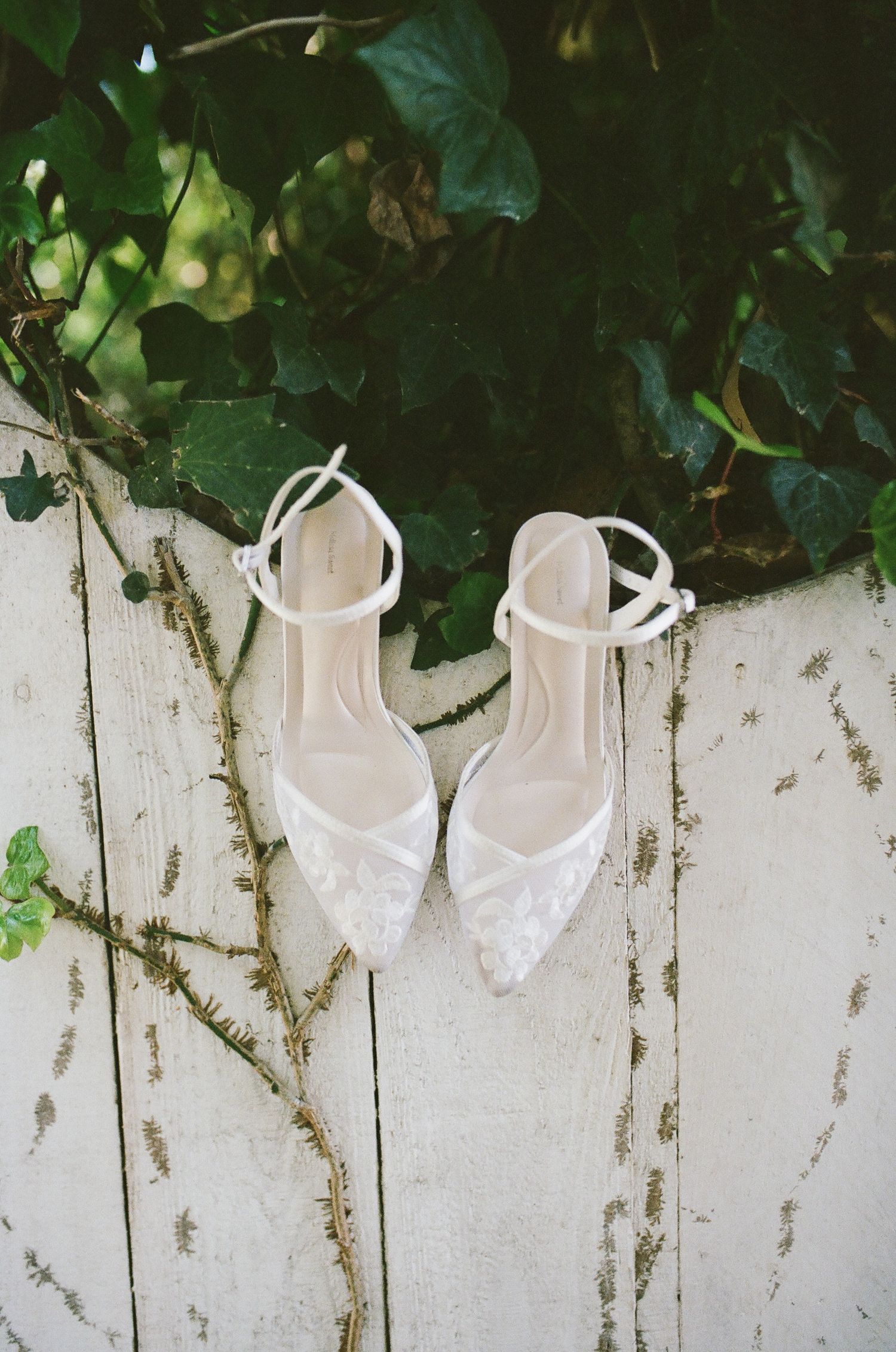 Ivory lace pointed-toe wedding heels with ankle straps resting on a weathered white wooden fence beneath green ivy leaves.