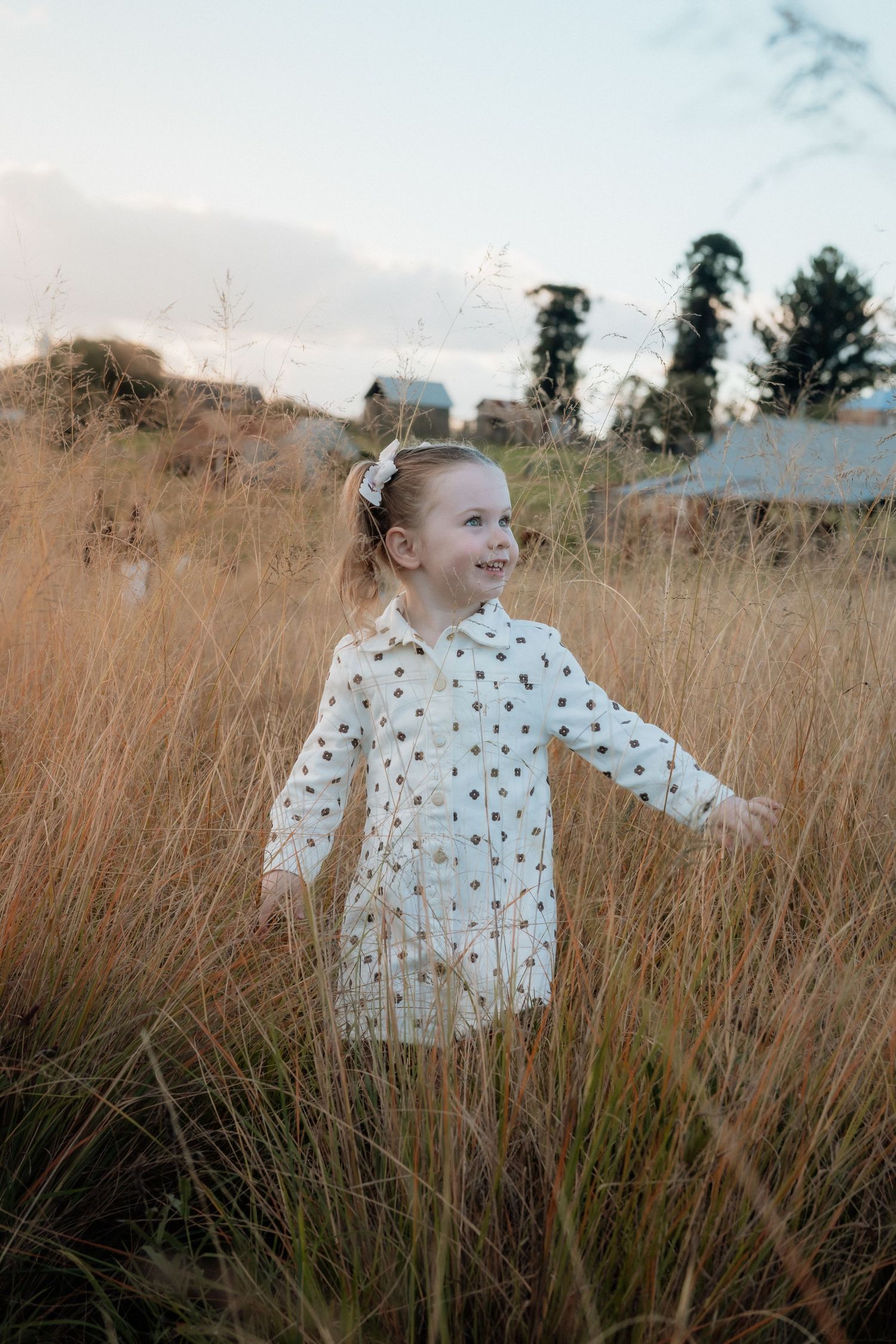 Smiling toddler girl in a white polka dot dress and hair bow stands with arms outstretched in tall golden grass at sunset in Rouse Hill.