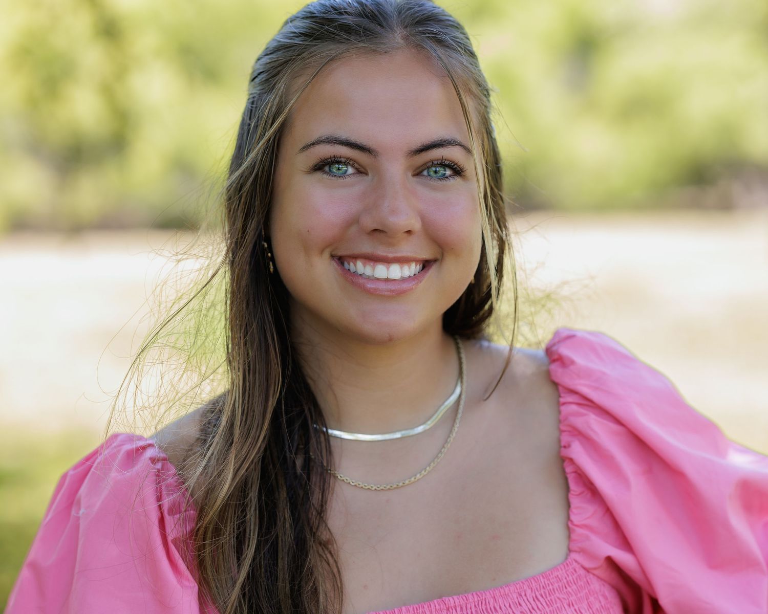 Portrait of someone in a pink dress with a pearl necklace smiling outdoors.