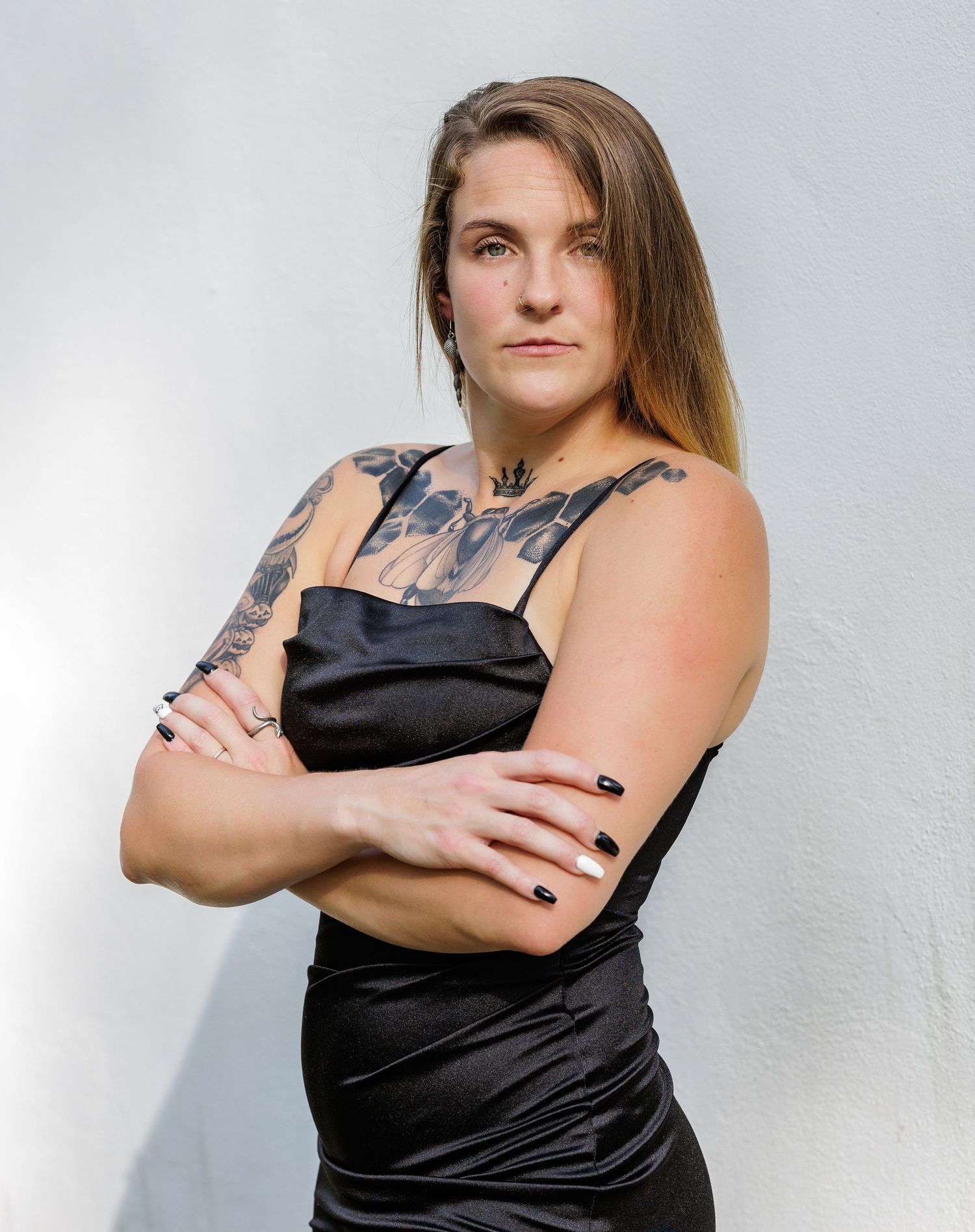 A person in a black dress poses confidently against a white wall, showing visible tattoo artwork.