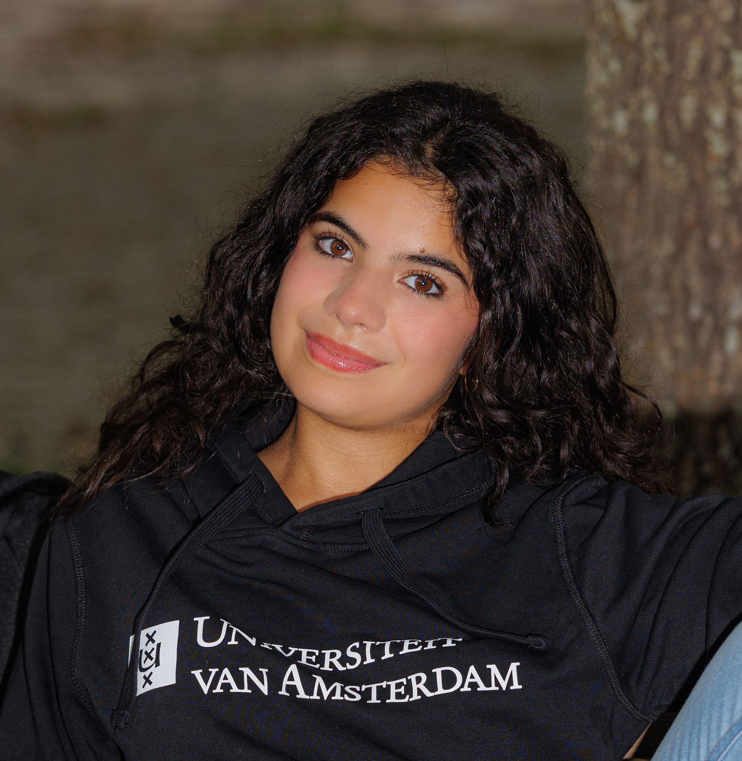 Young woman wearing a University of Amsterdam hoodie smiling outdoors with curly dark hair.