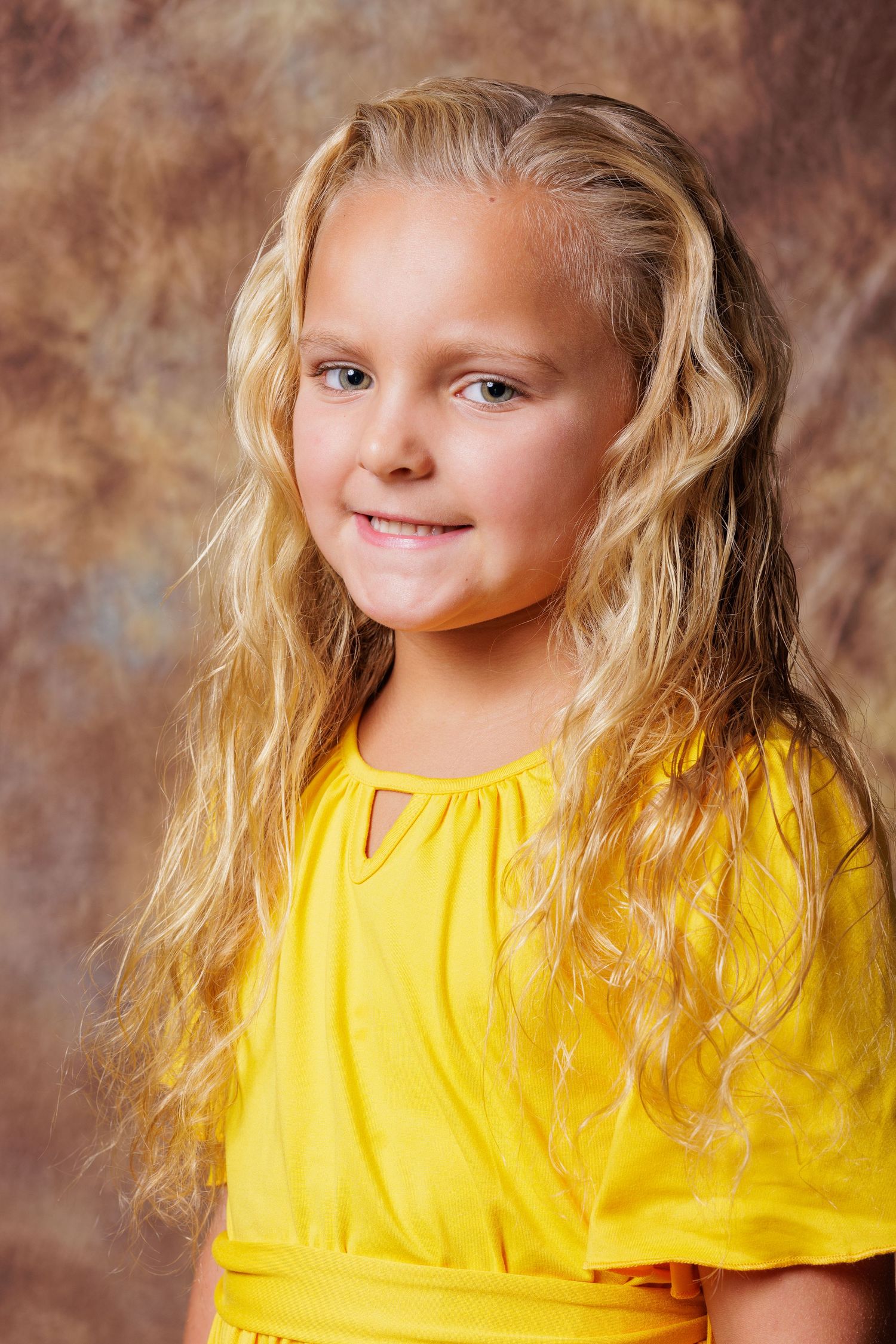 Young child wearing a bright yellow top smiling against a brown textured background.
