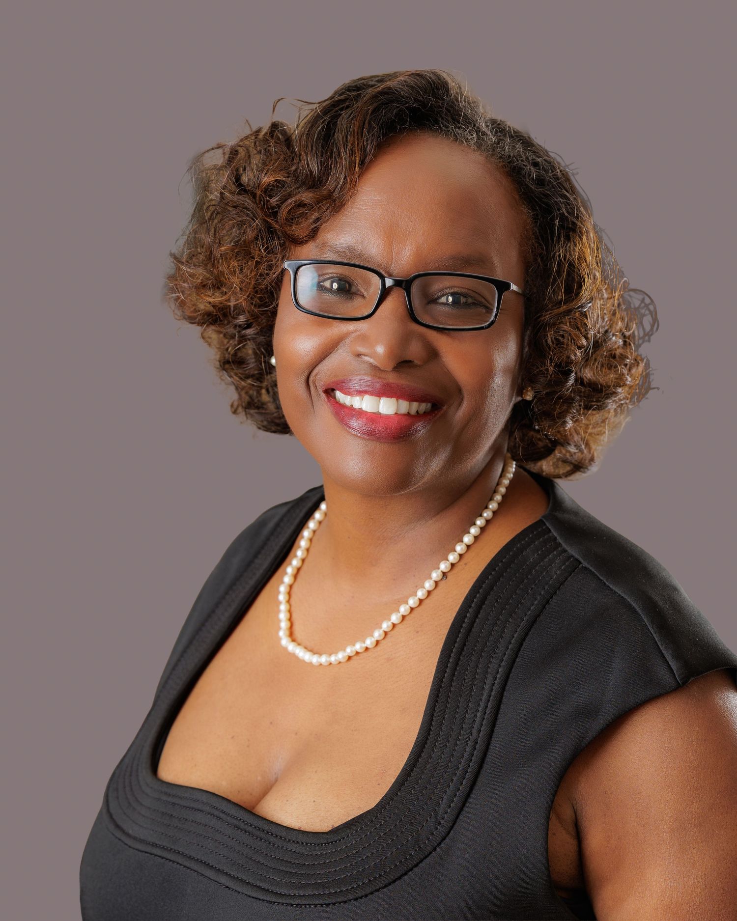 Professional portrait in black sleeveless top with pearl necklace and glasses against gray backdrop.