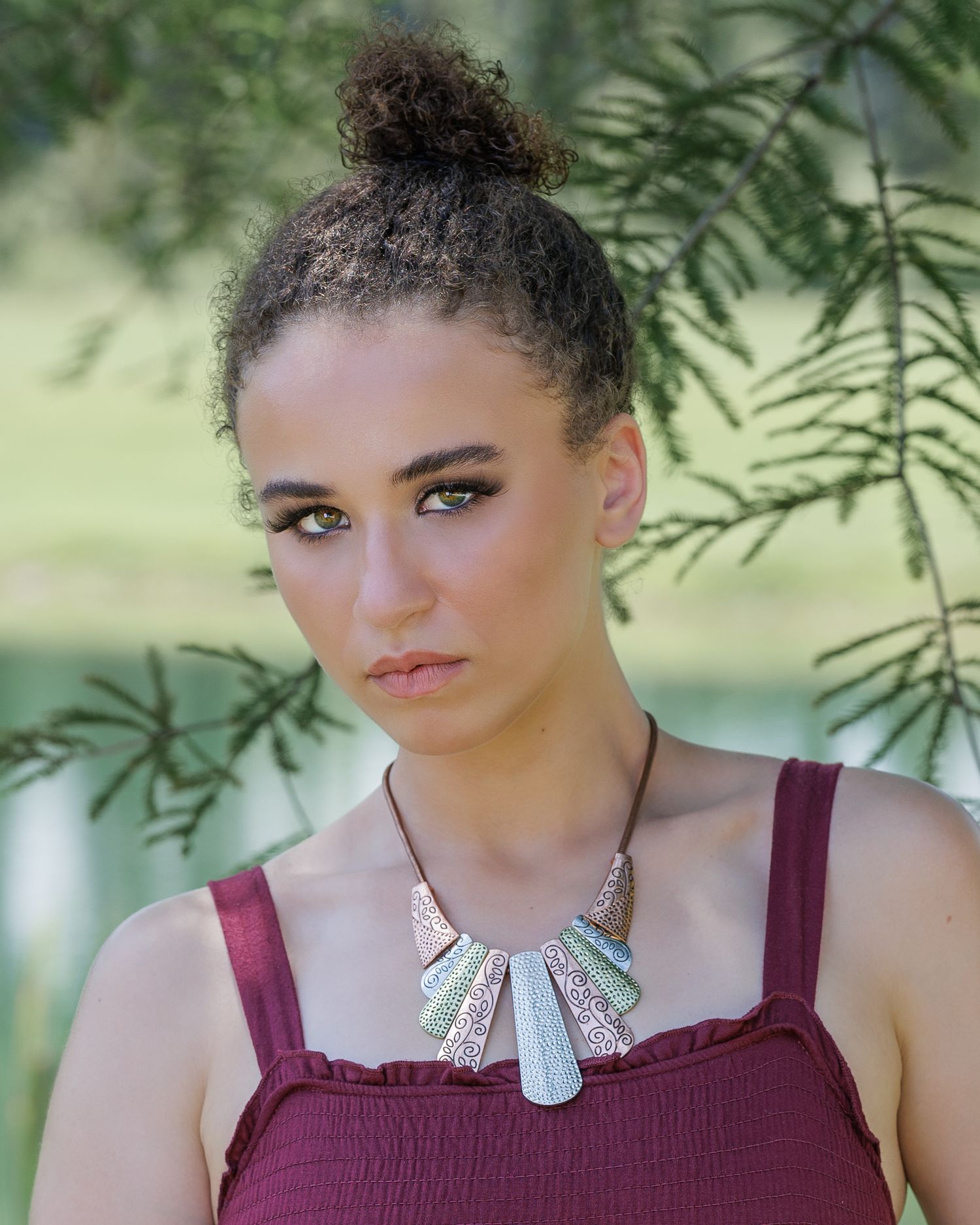 Young woman with curly updo wearing a burgundy top and silver statement necklace, posing outdoors near green foliage.