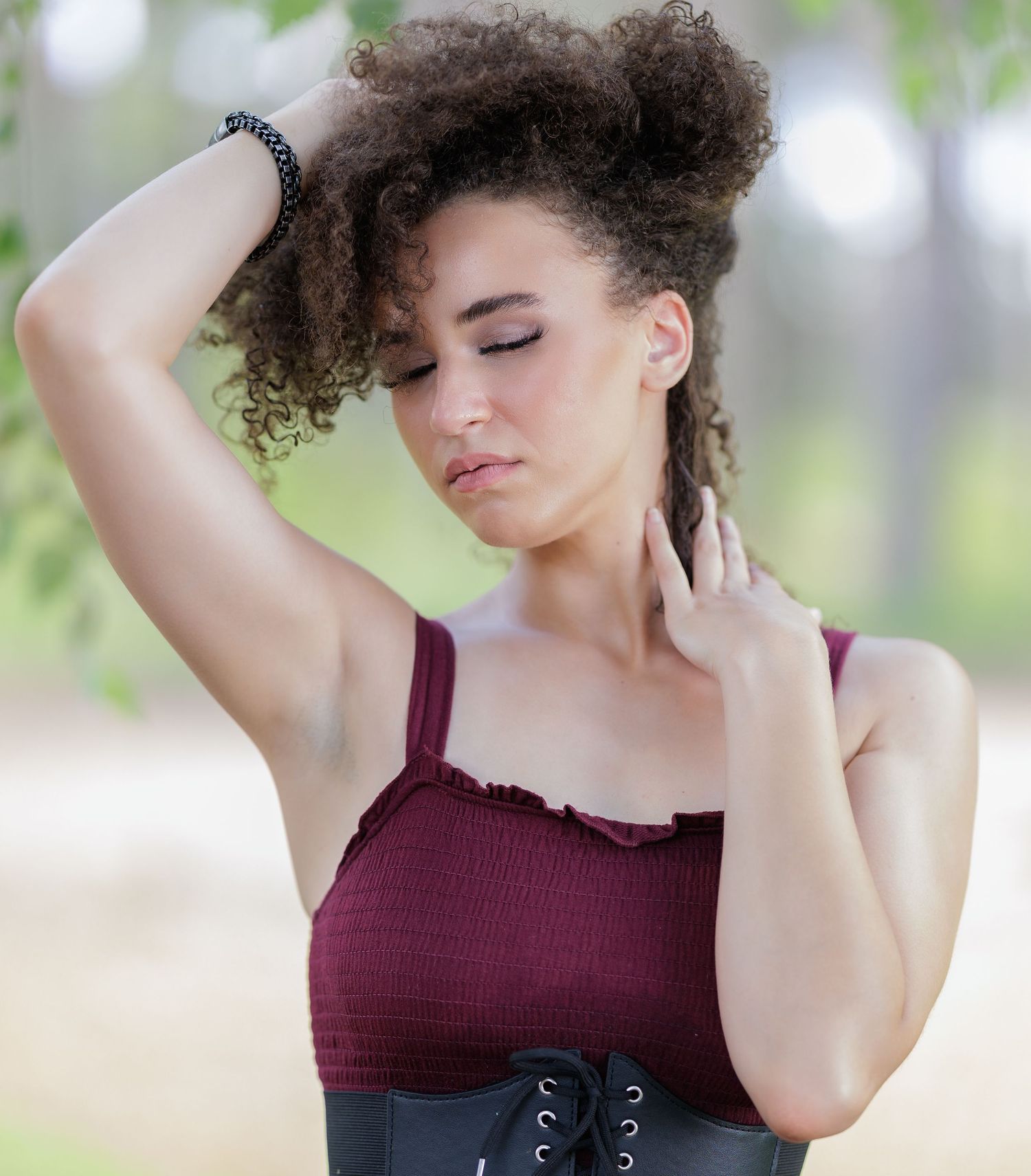 Young woman with curly hair in a burgundy crop top and black corset belt poses outdoors.