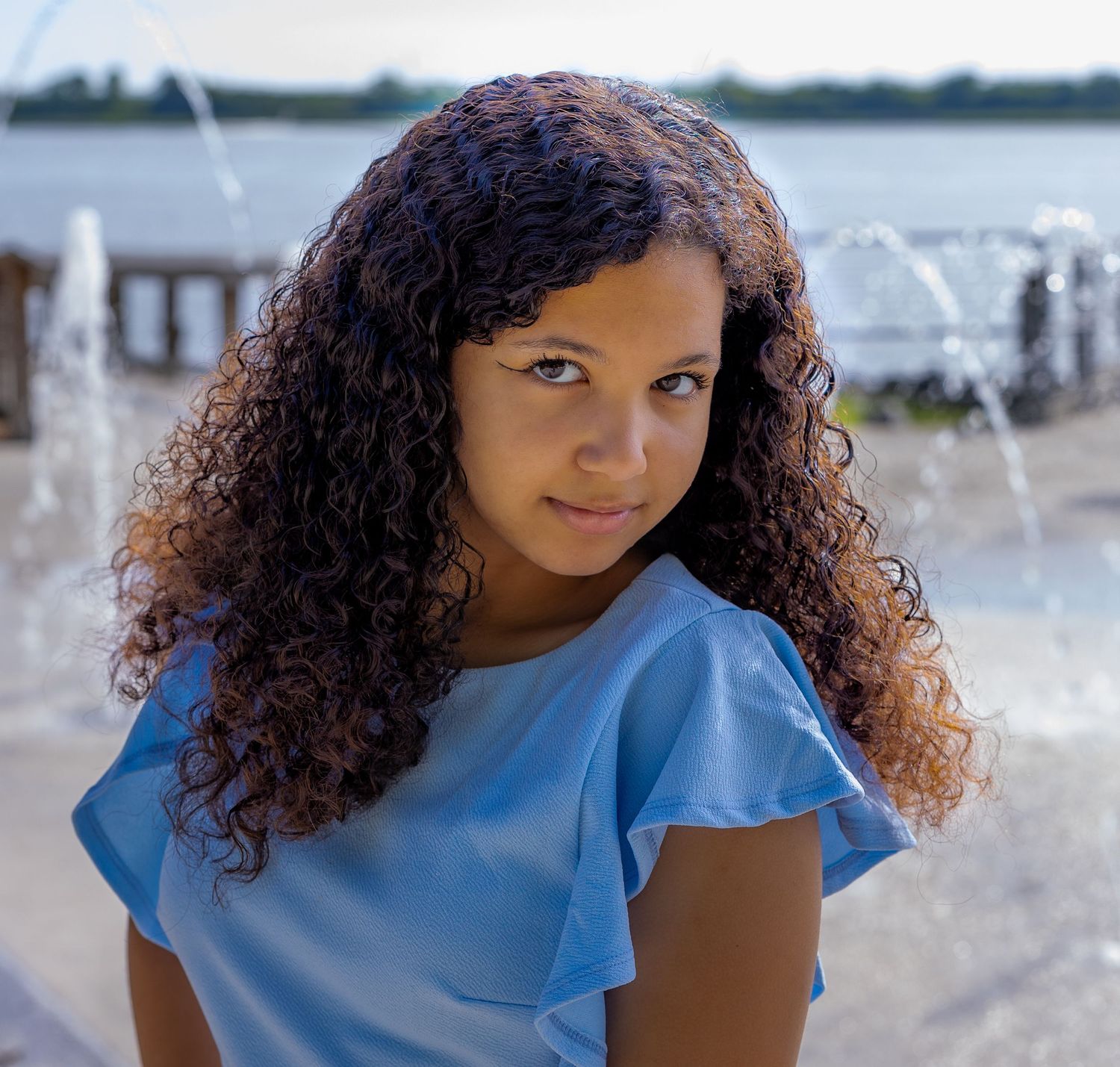 Portrait of a woman with curly hair wearing light blue top by a fountain.
