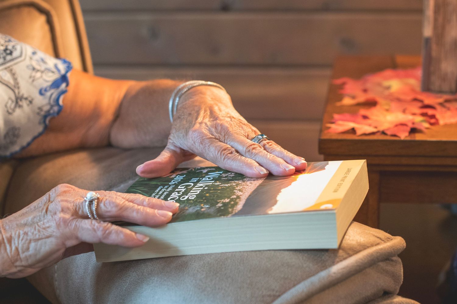 Elderly woman's hands with rings resting on an open book while relaxing in a cozy armchair.