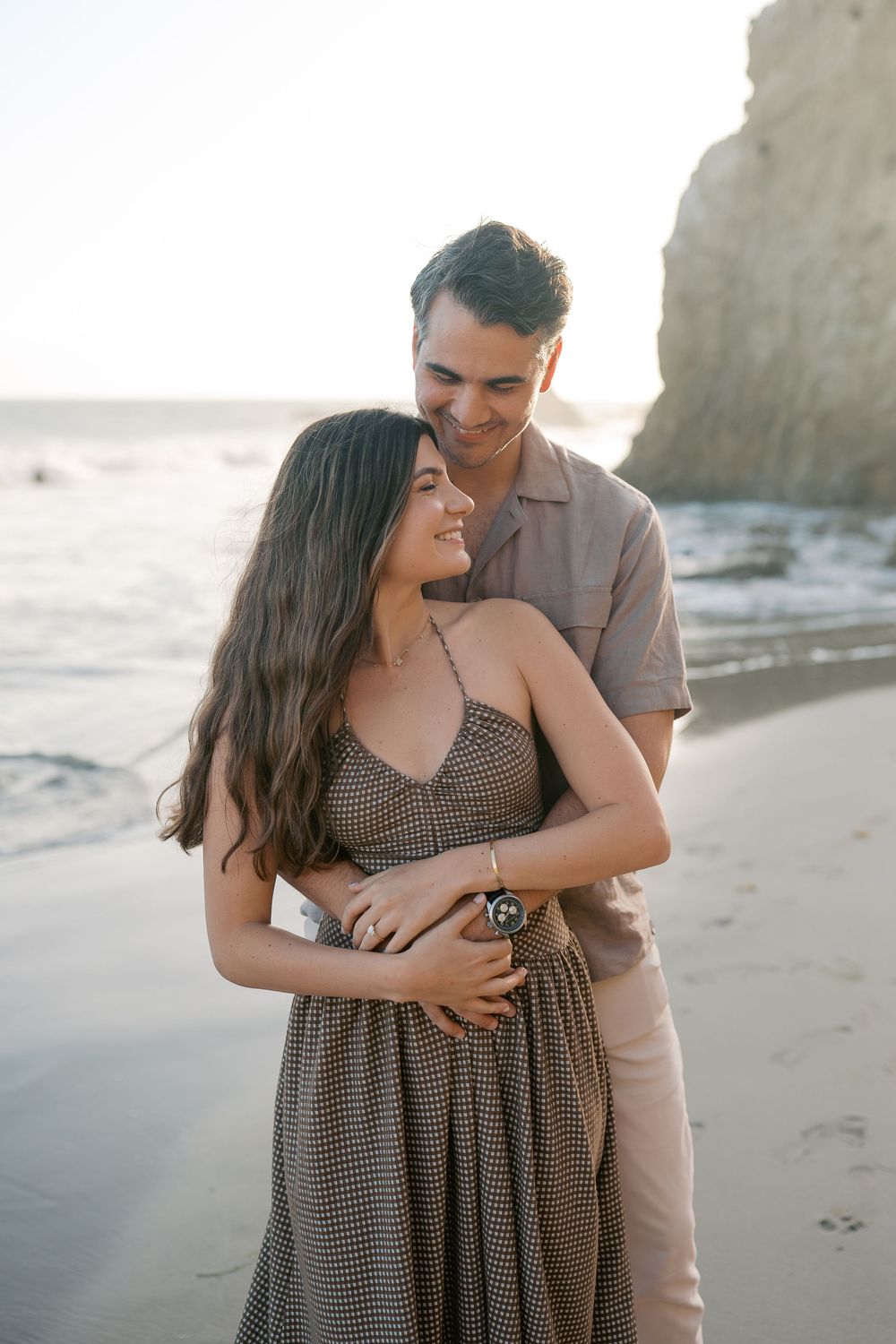 A couple embraces on a sandy beach at sunset, smiling warmly near ocean waves and a rocky cliff.