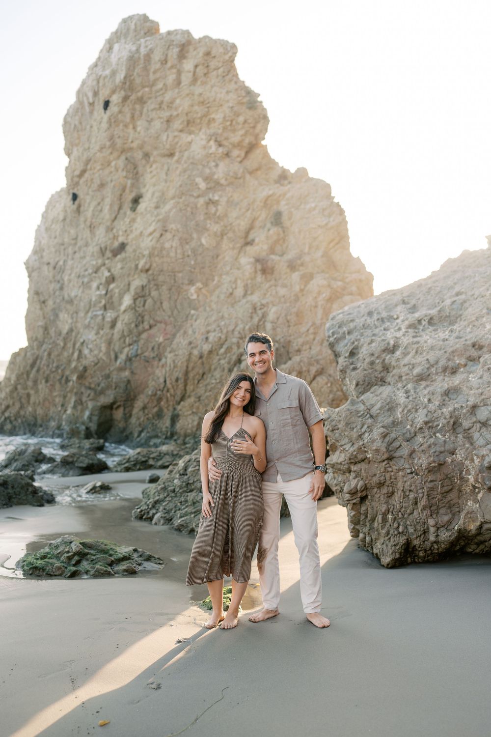 Couple posing barefoot on sandy beach with dramatic rock formations behind them at golden hour.