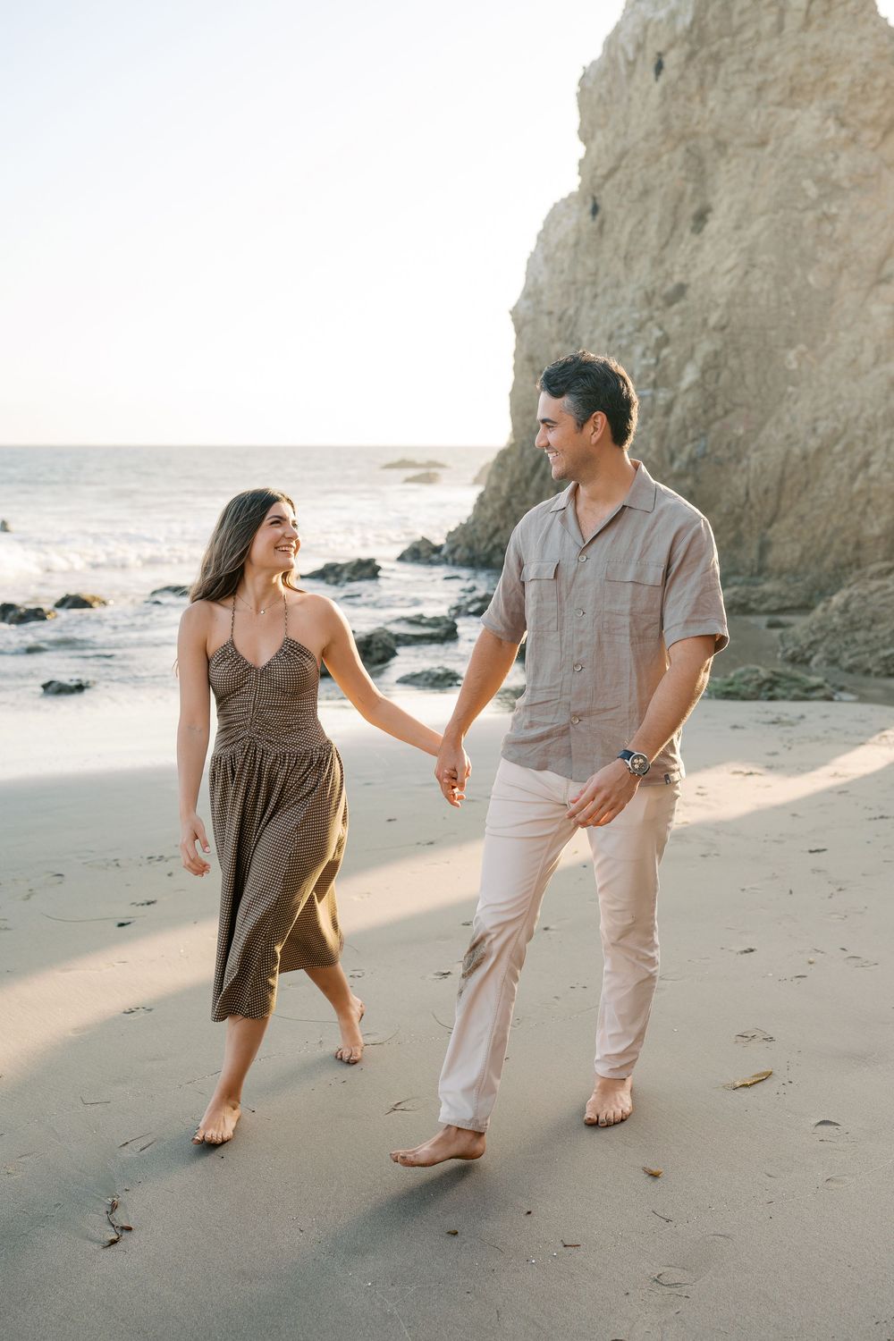 A couple holds hands and walks barefoot on a sandy beach near rocky cliffs, smiling at each other in casual summer outfits.
