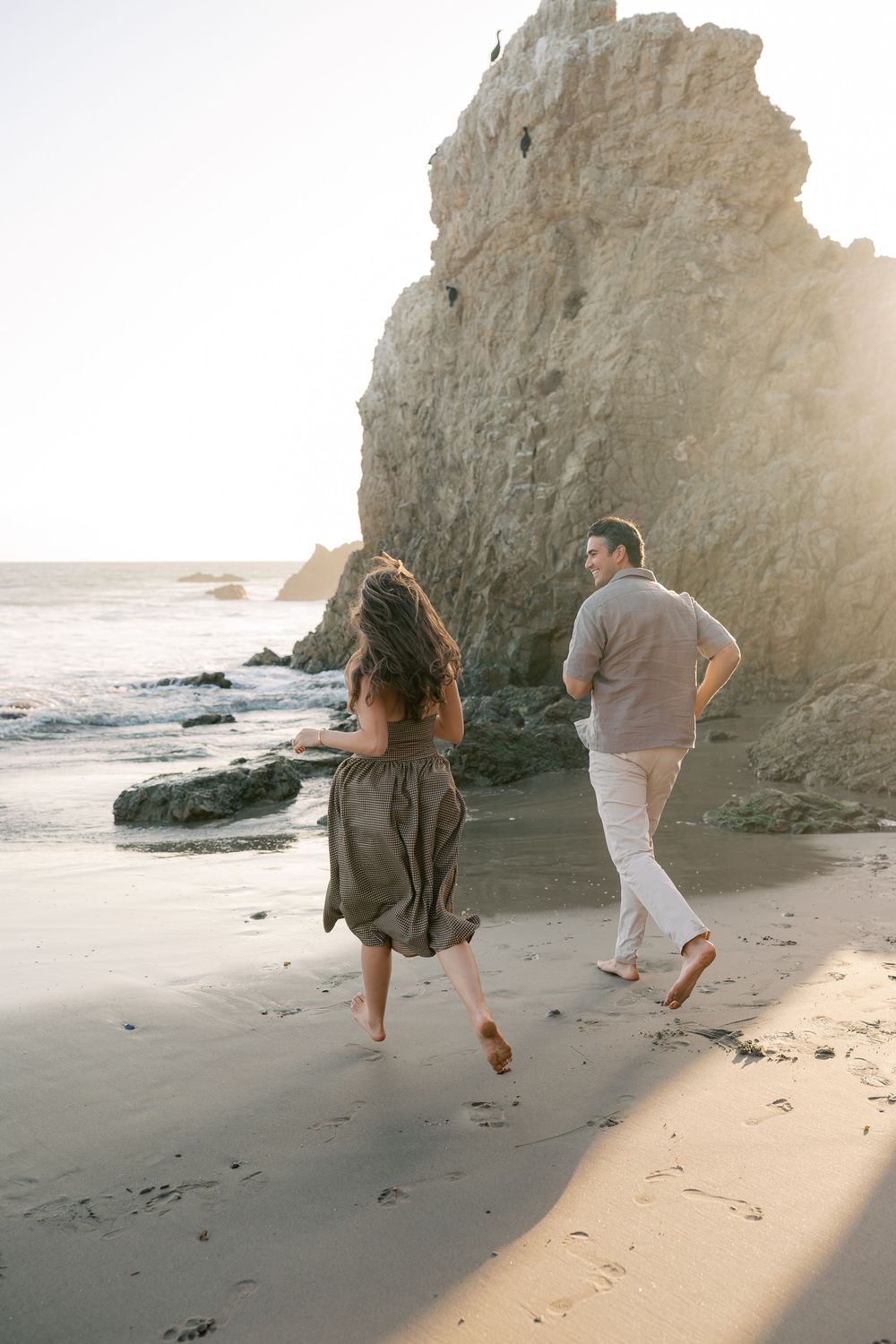 Joyful couple running barefoot along a scenic beach with dramatic rock formations at golden hour.