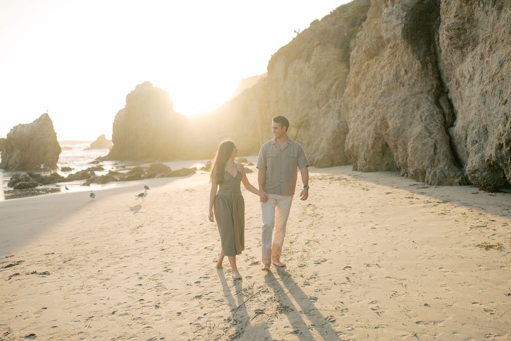 Couple holding hands walking on sandy beach at golden sunset with rocky cliffs in background.