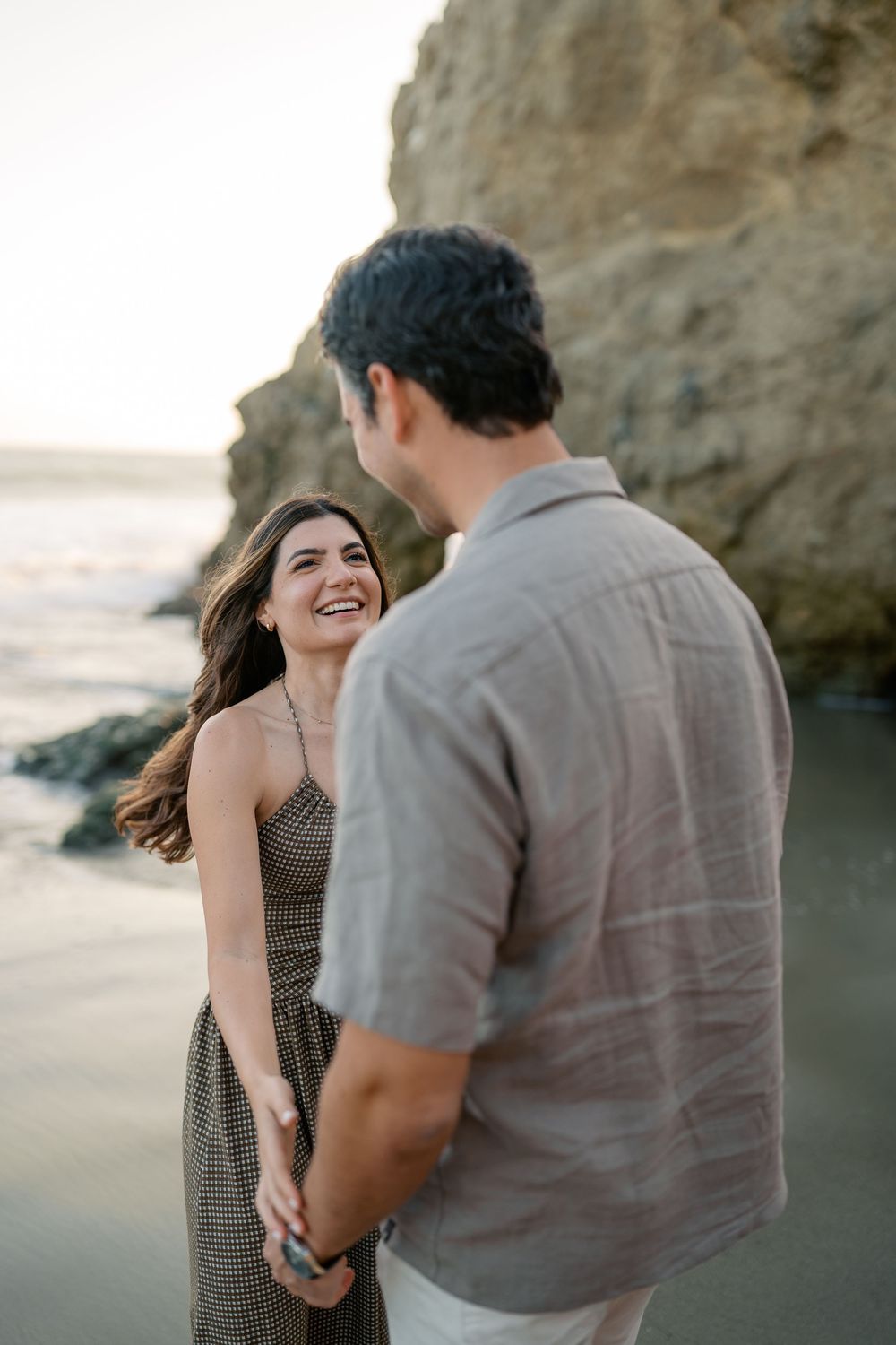 Happy woman laughing while holding hands with her partner on a rocky California beach at sunset.