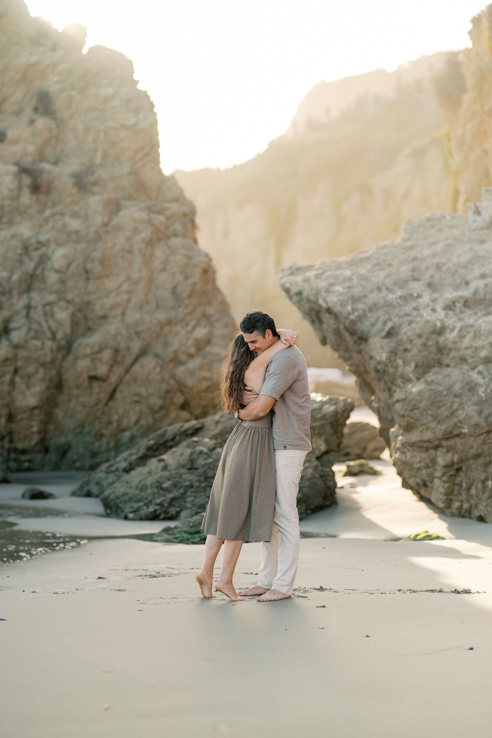 Couple sharing a tender embrace on a sandy beach surrounded by towering rock formations at sunset.