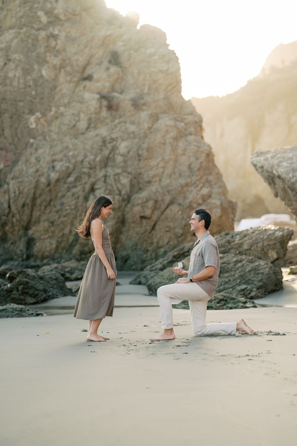 Man on one knee proposing to his girlfriend on a scenic beach with rocky cliffs at golden hour.