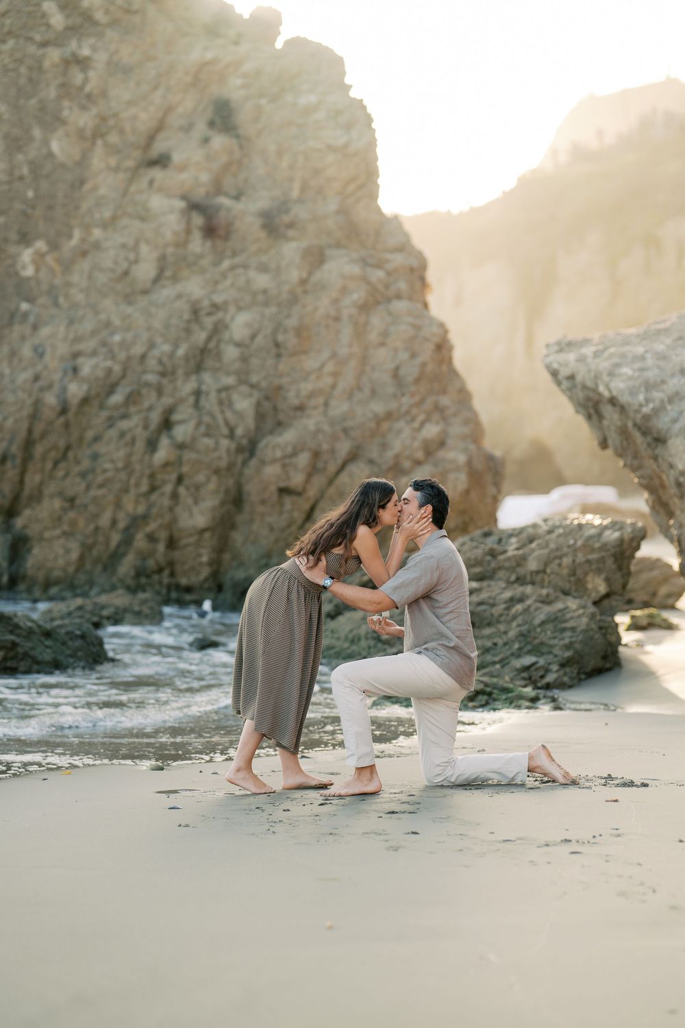 Man proposing on one knee to woman on sandy beach with rocky cliffs at sunset.