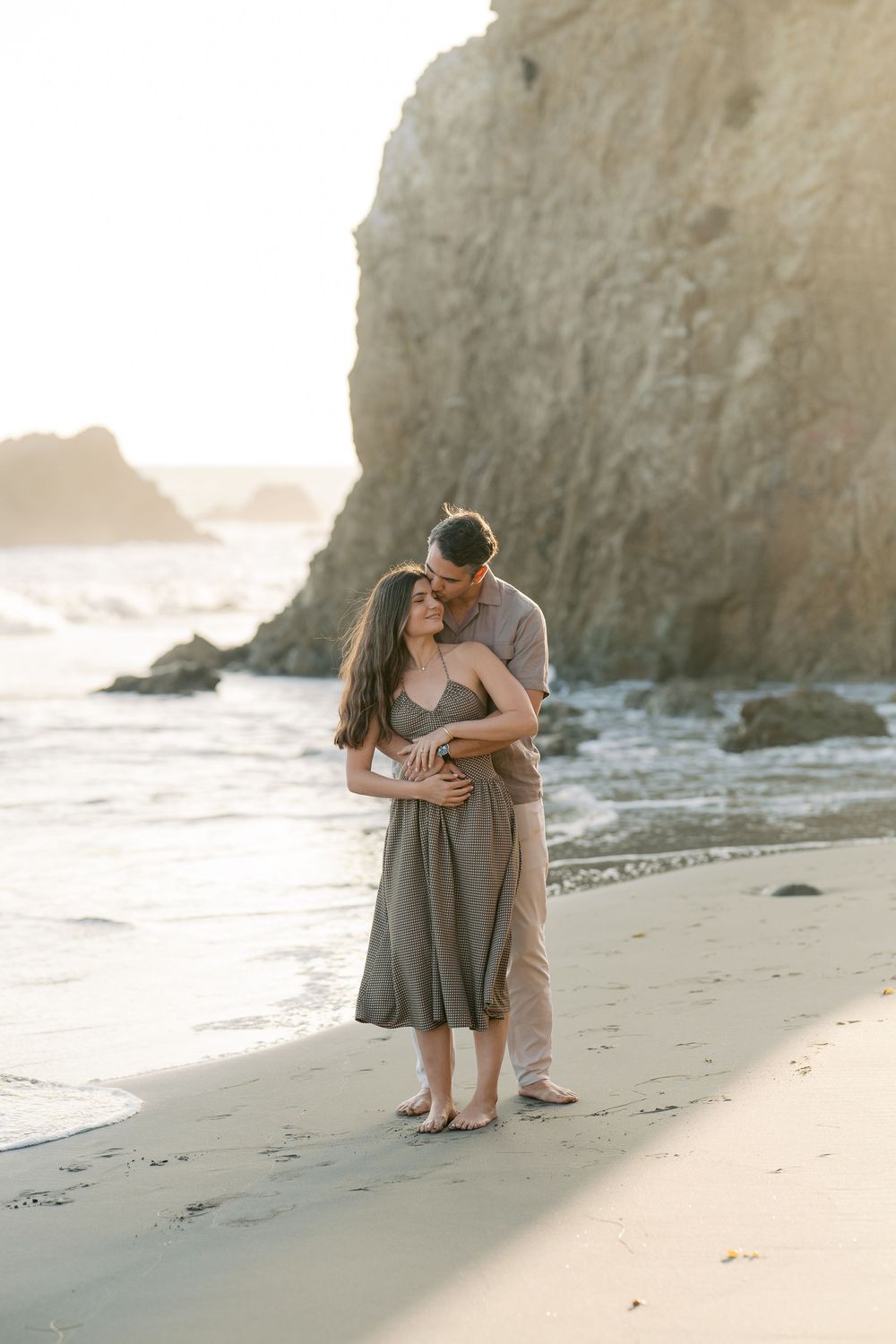 Couple sharing a tender embrace on a sandy beach with large rock formations at sunset.
