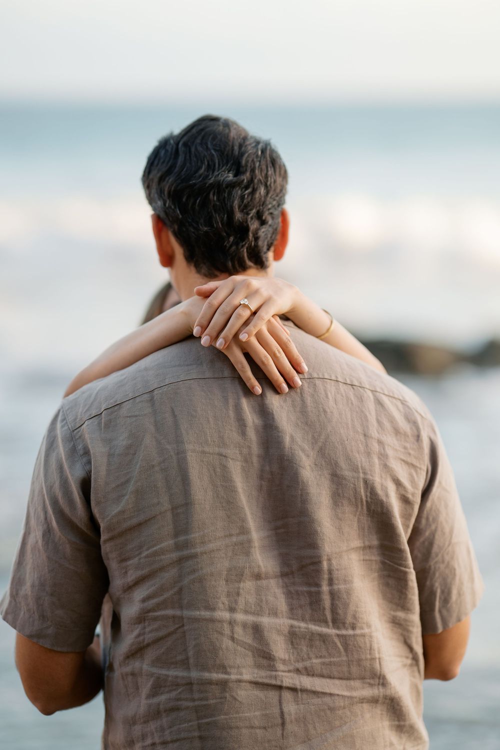 Couple embracing at the beach, woman's hand with engagement ring resting on man's shoulder, ocean in background.