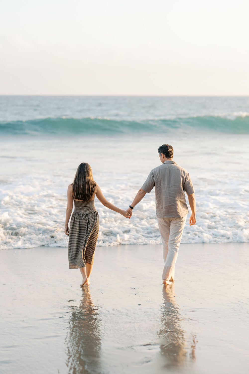 Romantic couple holding hands while walking toward ocean waves on a beautiful sandy beach at sunset.