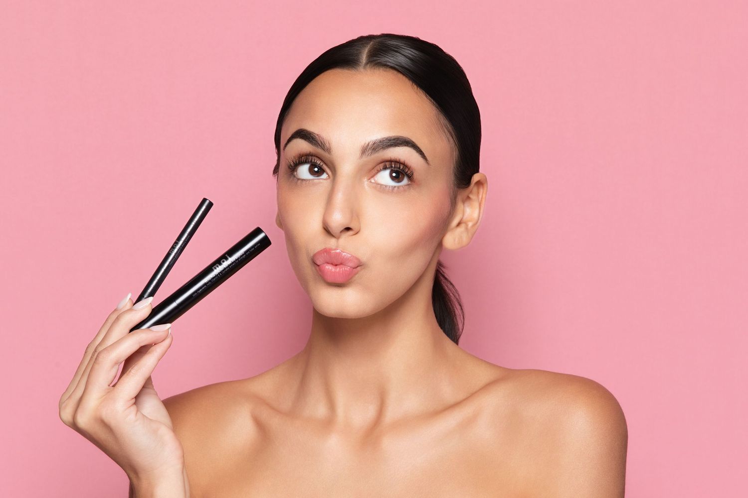 Woman holding makeup brushes with pouty lips against pink background, showcasing beauty cosmetics tools.