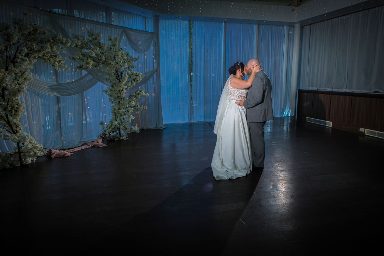 Bride and groom share first dance in elegant blue-lit wedding venue with floral arch backdrop.