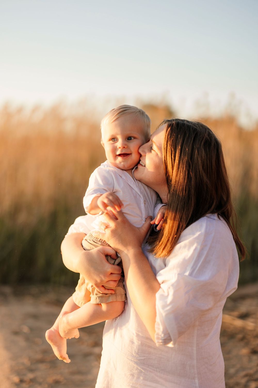 Mother kissing happy baby on cheek while holding her in sunny wheat field.