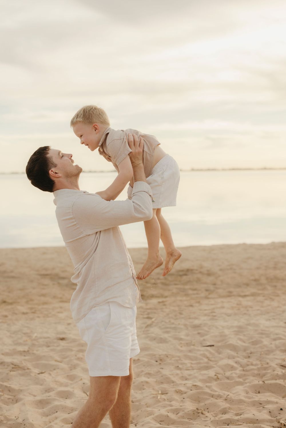 Father lifting smiling toddler high in the air on sandy beach at sunset.