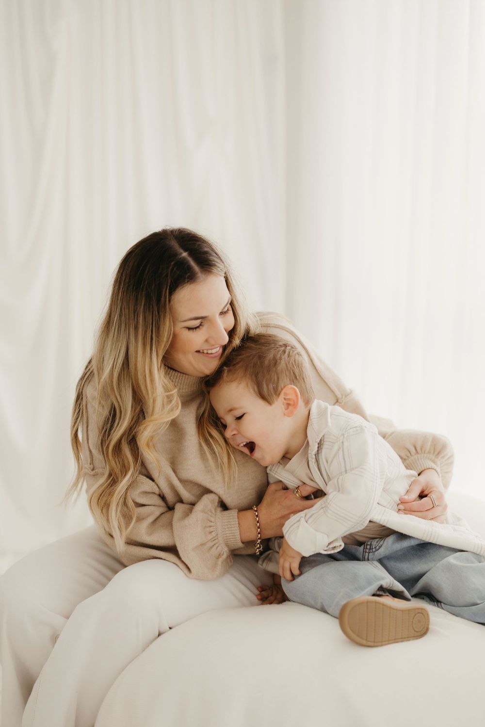 A smiling mother cuddles her laughing toddler on a white sofa in a bright, airy room.
