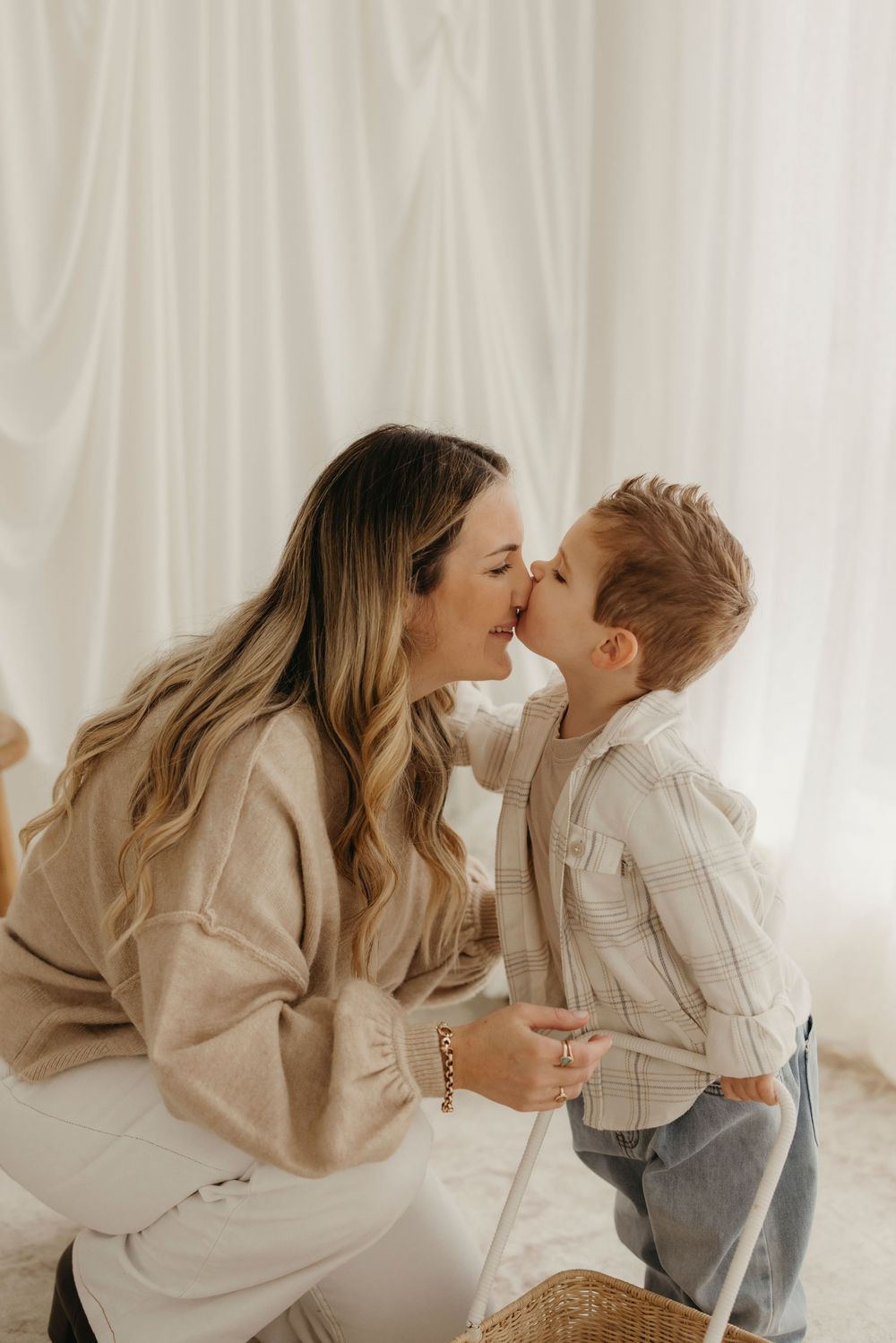 A loving mother shares a sweet kiss with her young son in a bright white studio setting.