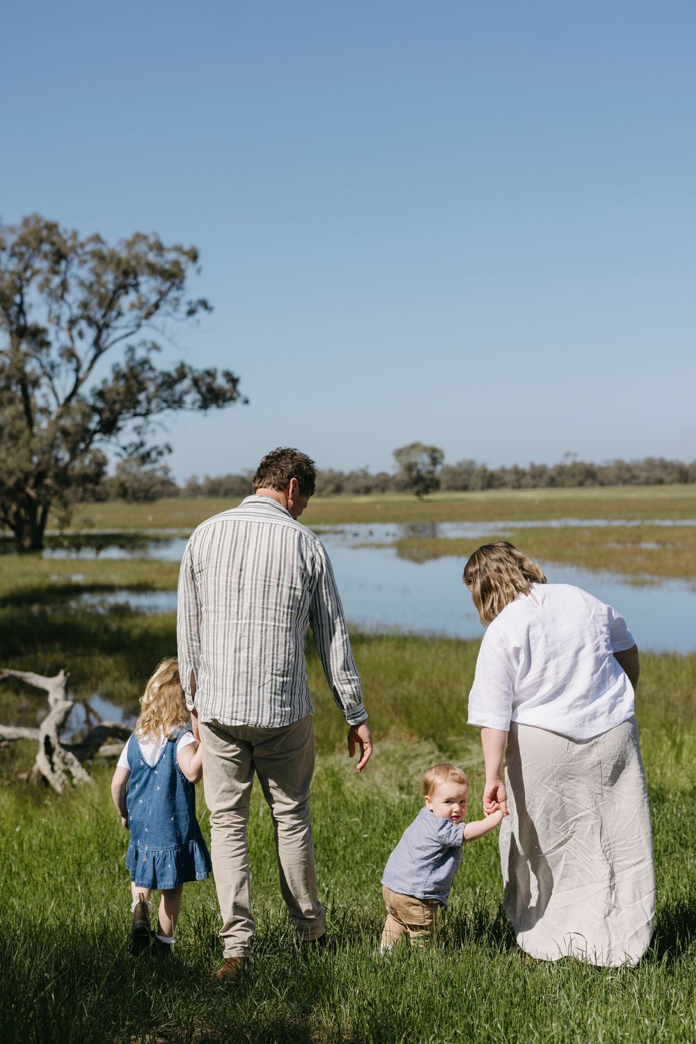 A family of four walks together through a lush green field beside a tranquil pond at sunset.
