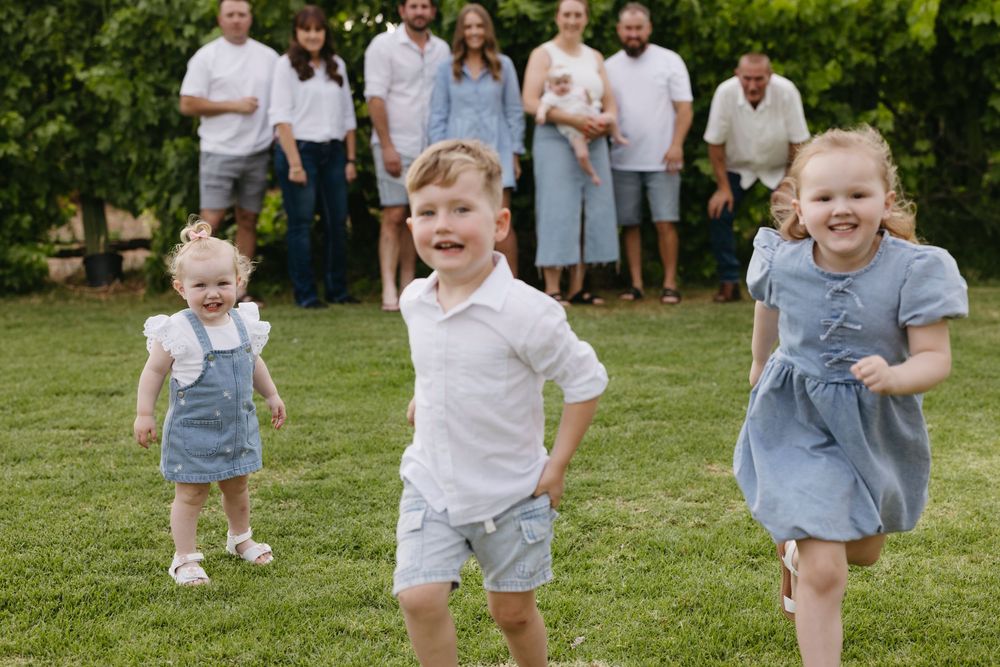 Three young children running toward camera with family group standing on green lawn.